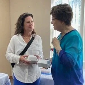 Two women having a conversation indoors, one holding a white box and a paper, the other gesturing with her hand, in front of a window with patterned curtains.