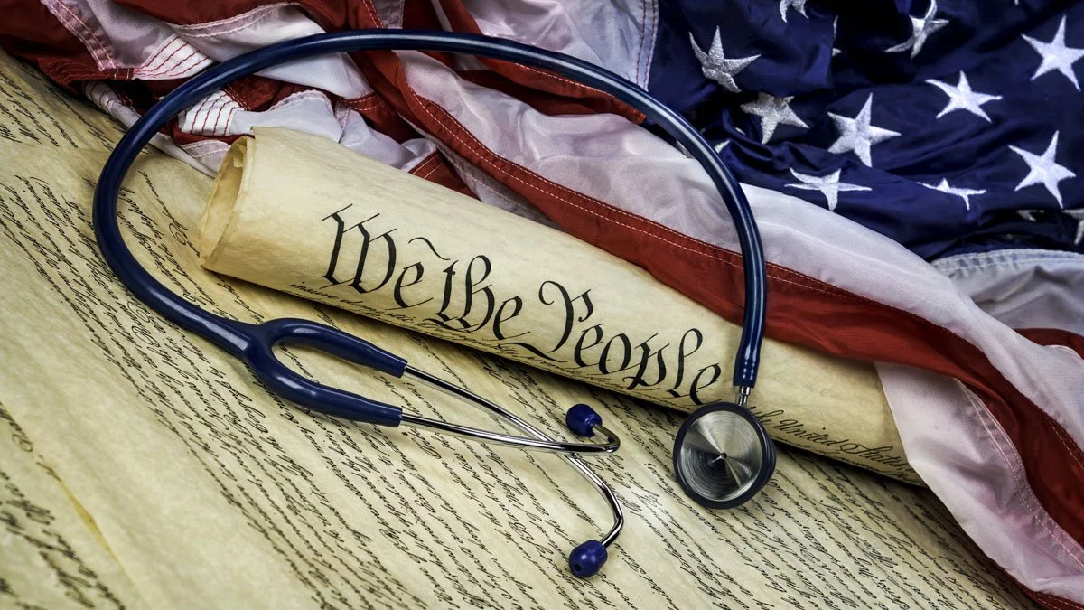 A stethoscope resting on a rolled-up copy of the US Constitution, with an American flag in the background.