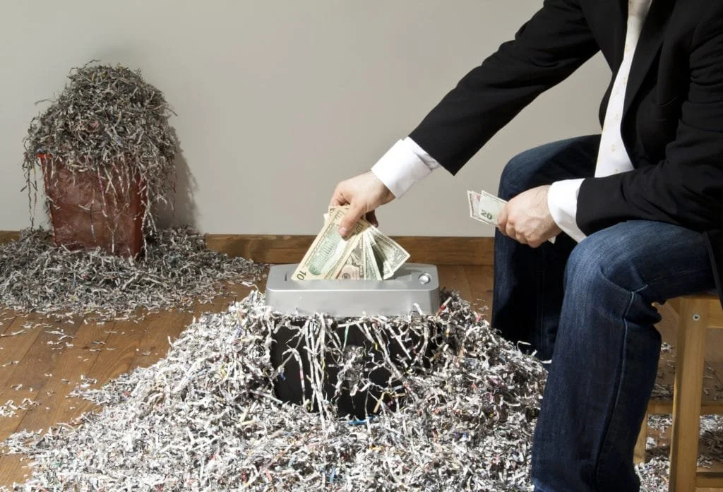 Person in a black blazer and jeans putting money into a shredder surrounded by shredded paper and confetti on the floor, with a potted plant covered in shredded paper in the background.