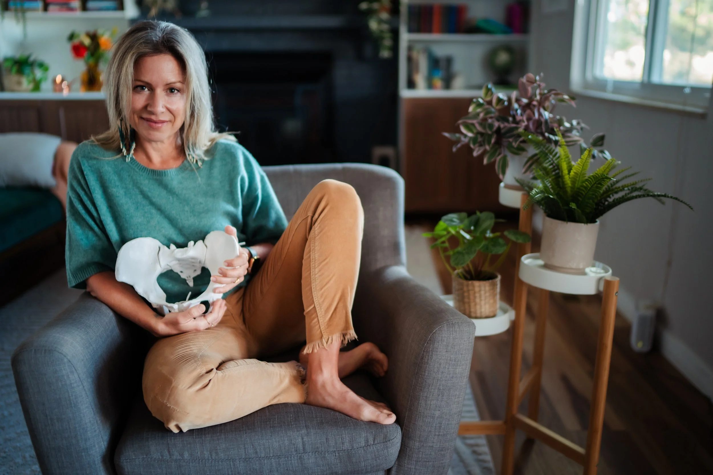 A woman sitting on a gray armchair in a living room, holding a model of a pelvis and smiling. The room is decorated with potted plants and has a large window with natural light.