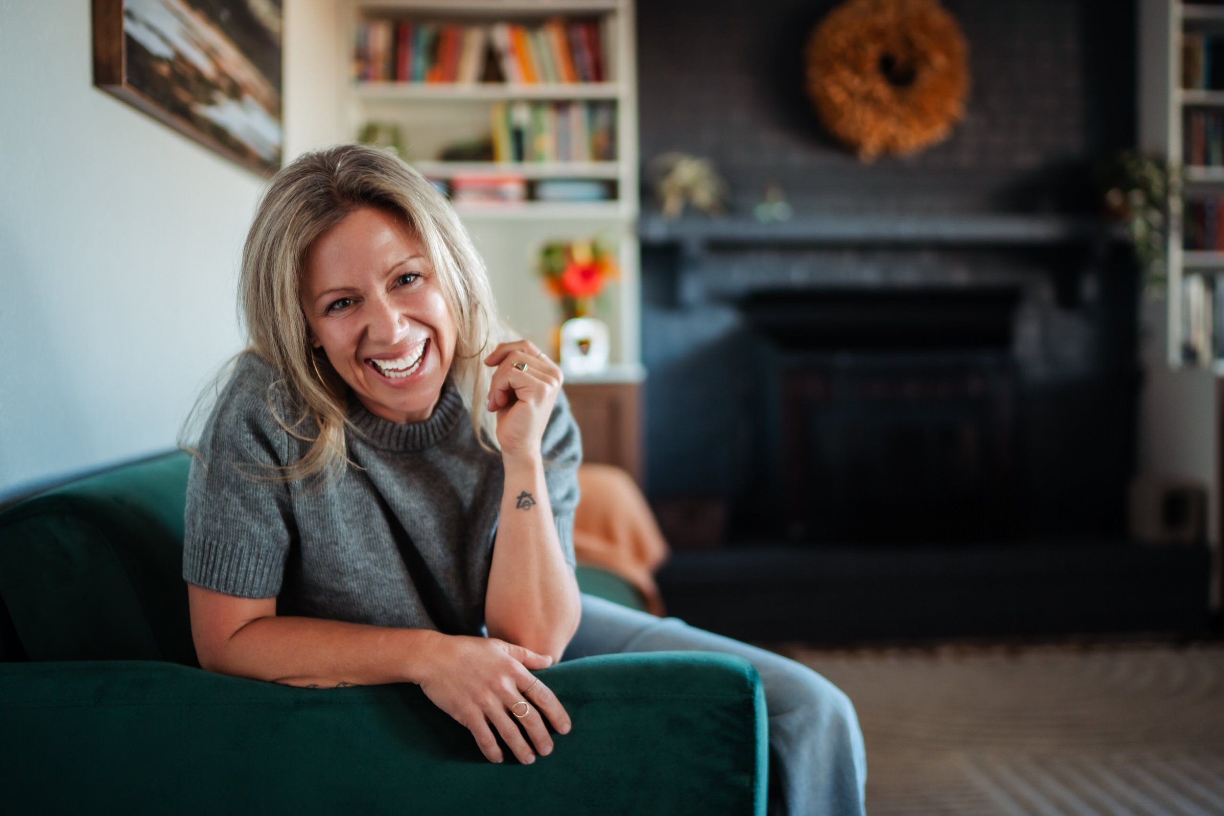 A woman with blonde hair smiling and laughing while sitting on a green couch in a cozy living room with bookshelf, fireplace, and decorative items in the background.