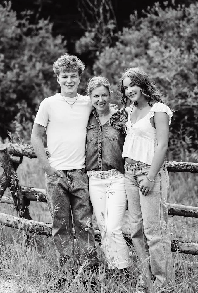 Black and white photo of a happy woman with two teenagers, a boy on her left and a girl on her right, standing outdoors with a rustic wooden fence and trees in the background.