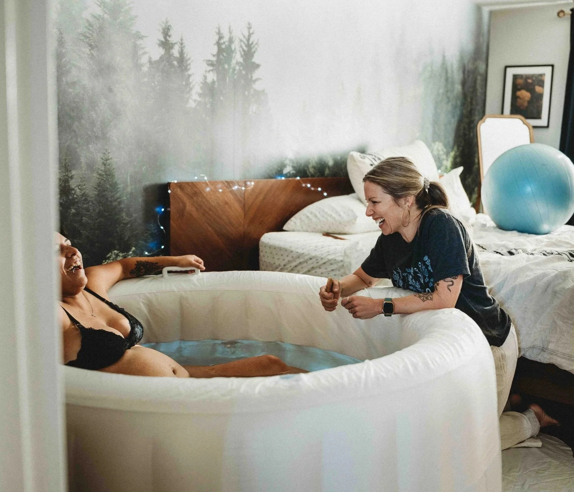 Two women sharing a laugh in a bedroom, with one woman sitting in a small inflatable pool and the other leaning over the edge. The woman in the pool is wearing a black bra, and the woman leaning is wearing a black T-shirt and beige pants. The room has a nature-themed wall mural, a bed with a large blue exercise ball on it, fairy lights, and a framed picture on the wall.