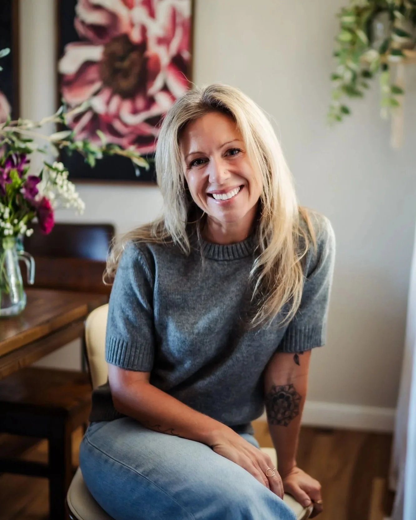 A woman with long blonde hair, wearing a gray sweater, smiling and sitting on a chair in a room with a floral painting and a table with a vase of flowers.