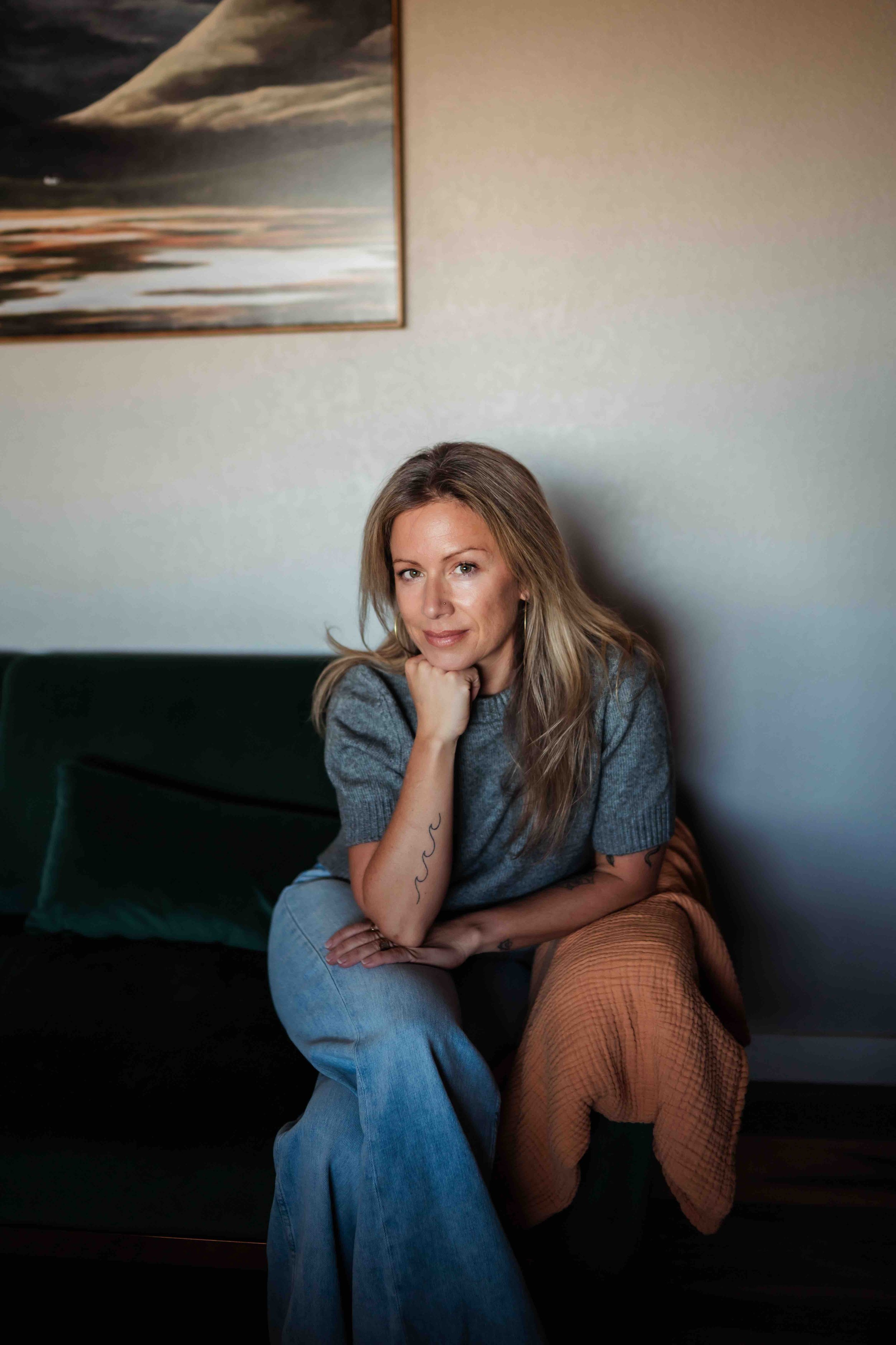 A woman with blonde hair sitting on a dark green couch, resting her chin on her hand, wearing a gray top and blue jeans, with a textured orange blanket draped on the side of the couch, in a room with a beige wall and a landscape painting.