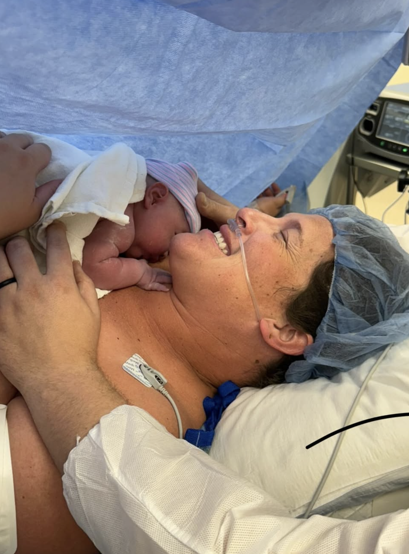 A woman in hospital attire and a shower cap is lying in a hospital bed, smiling with her eyes closed, as she holds her newborn baby close after childbirth, with a nasal oxygen tube attached.