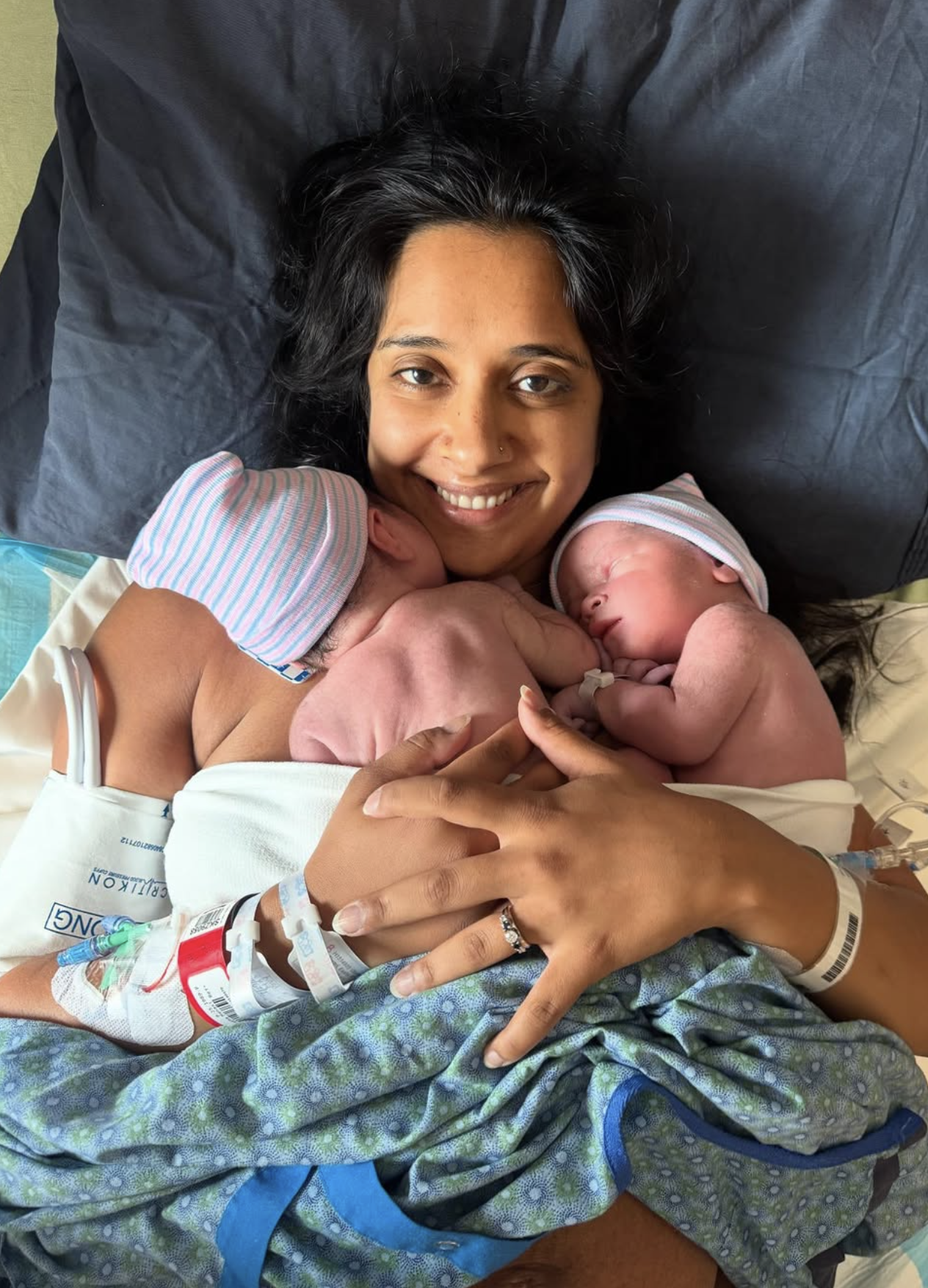 A woman lying in a hospital bed after childbirth, holding her newborn twins who are breastfeeding, wearing hospital caps and with medical tubes and bracelets visible.