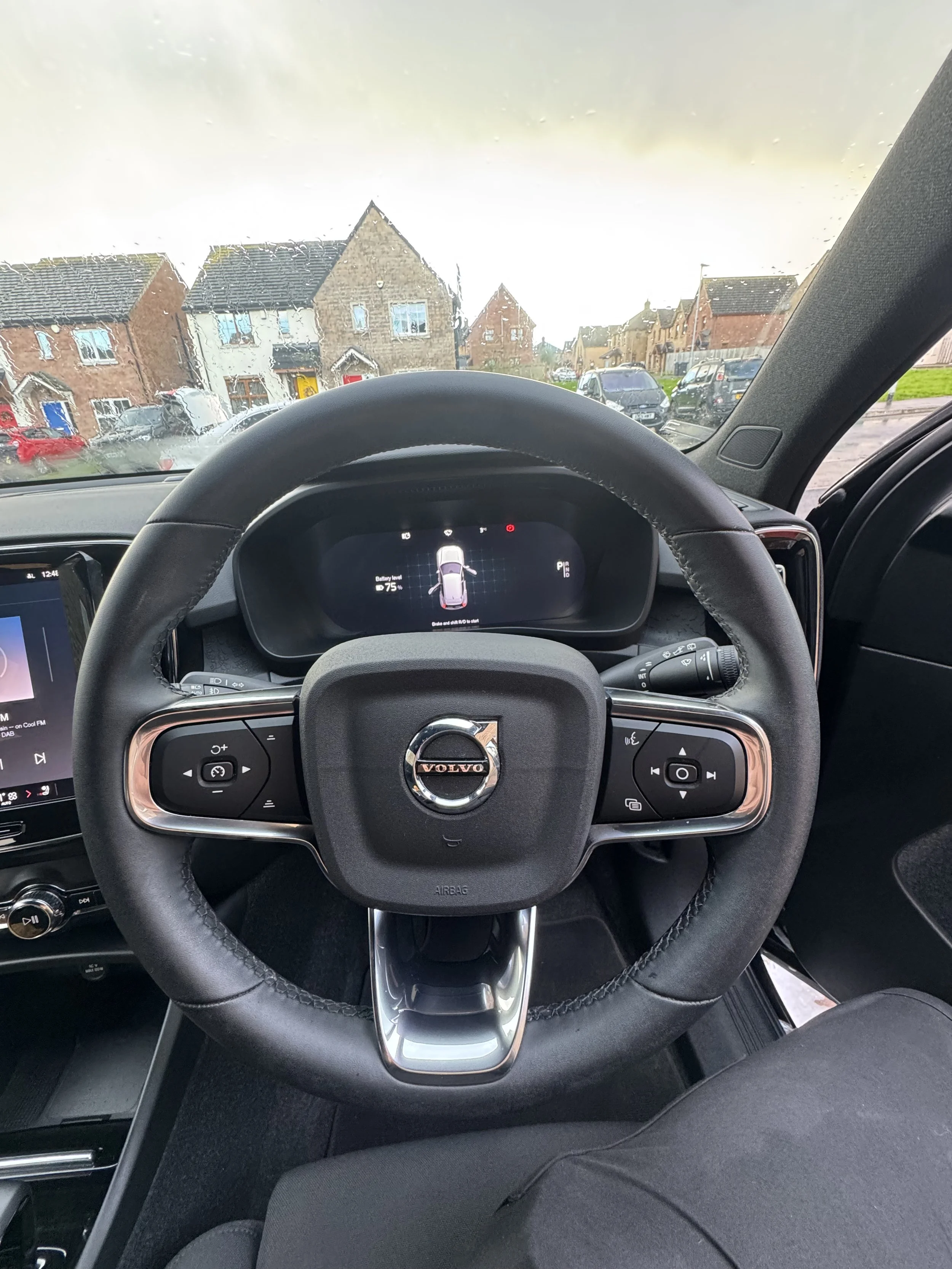 Inside view of a Volvo car dashboard, showing a steering wheel with control buttons, digital instrument panel, and part of an infotainment screen, with houses visible through the windshield on a rainy day.