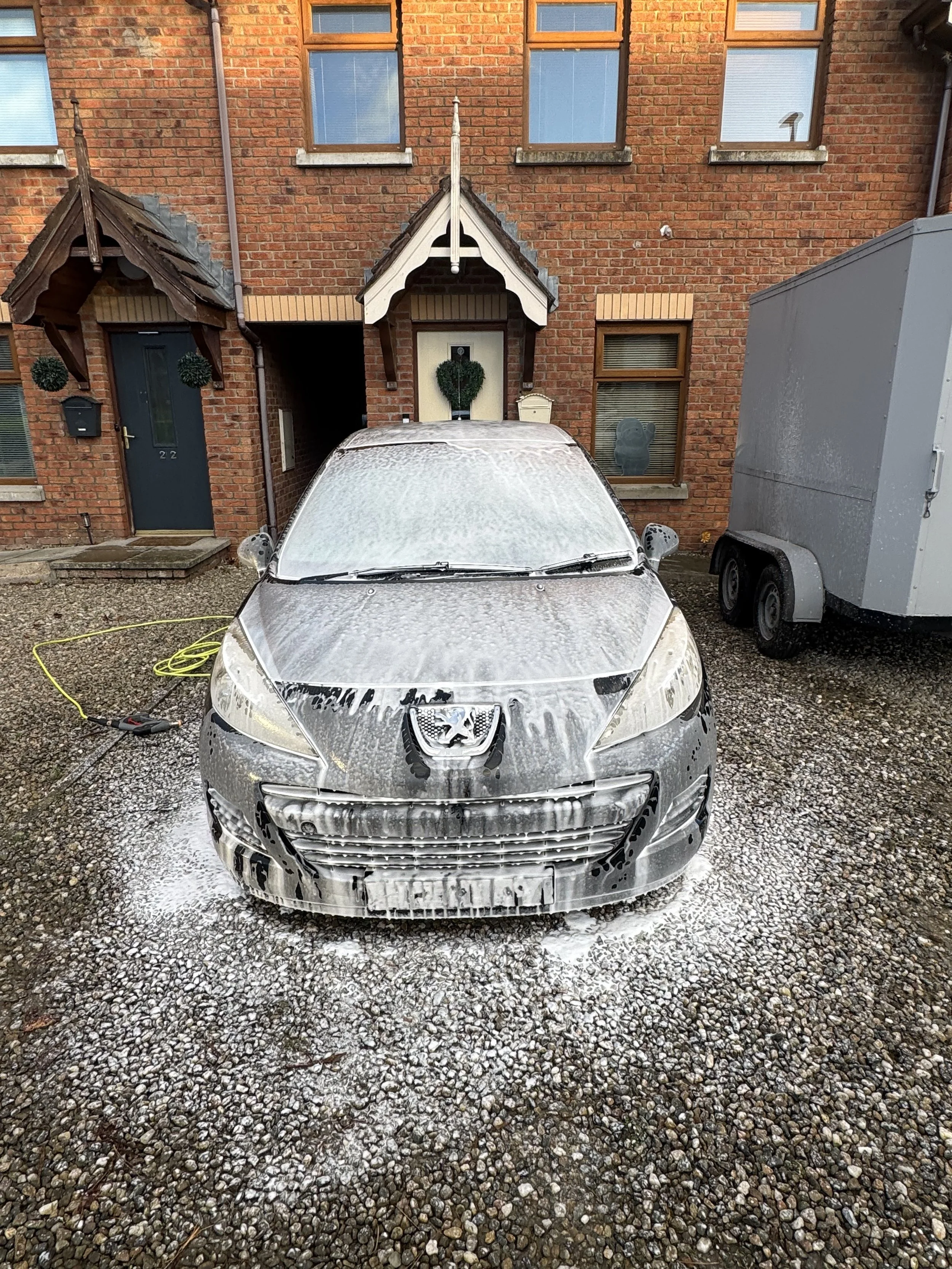 A grey Peugeot car being washed with foam, parked on a gravel driveway outside a brick house with three windows and two front door canopies.
