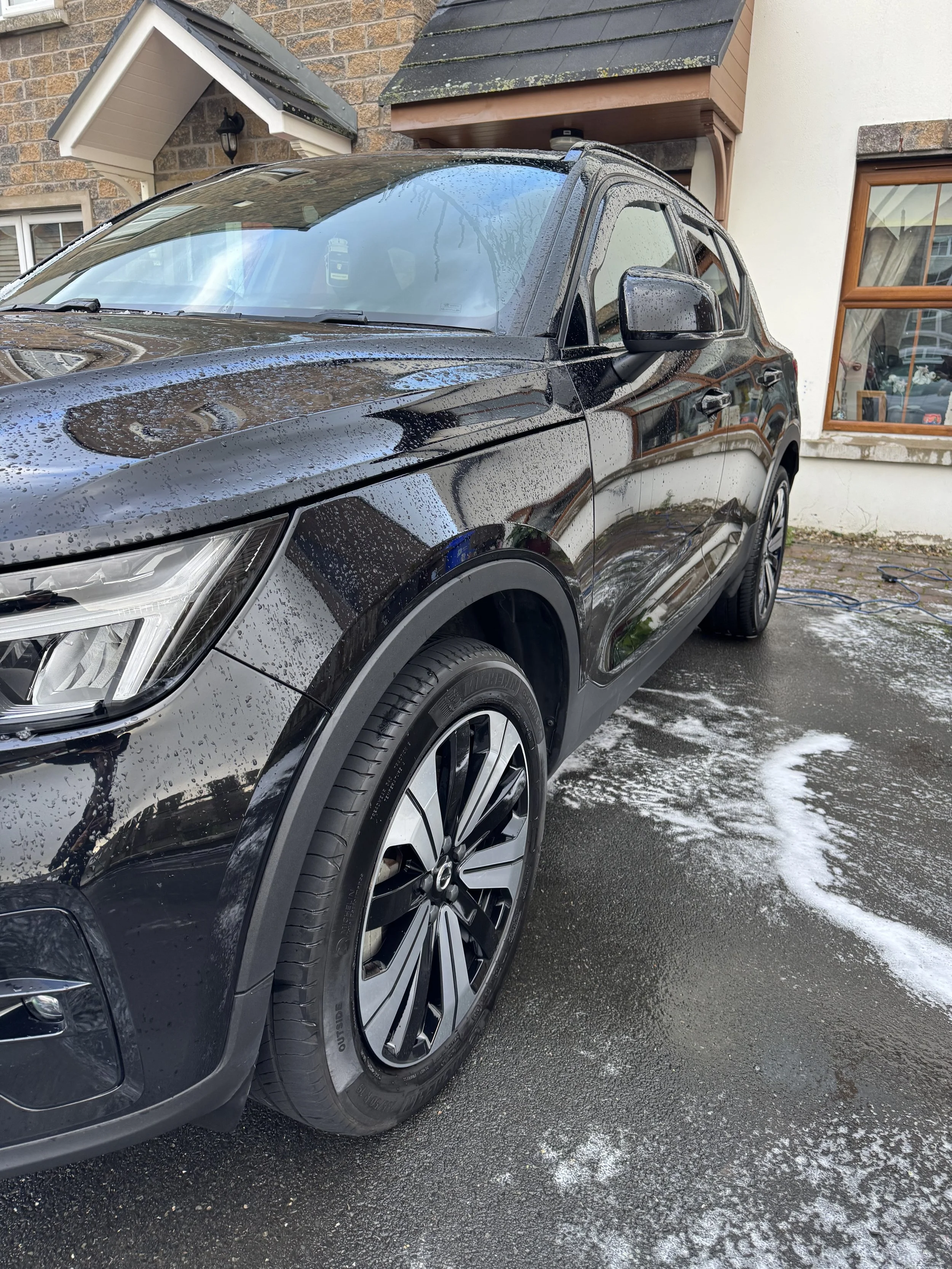 Black SUV parked outside a house with wet ground and soap suds from washing the car, reflecting a house and trees.