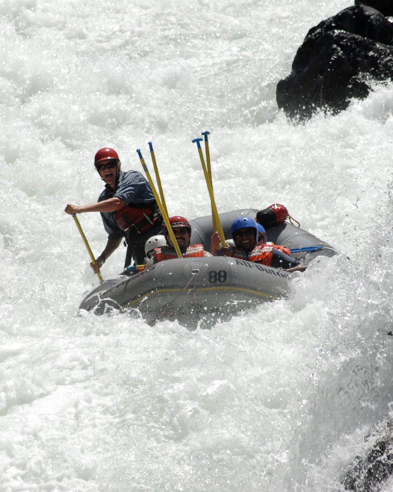 People white-water rafting on a river with waves and rocks, wearing life jackets and helmets.