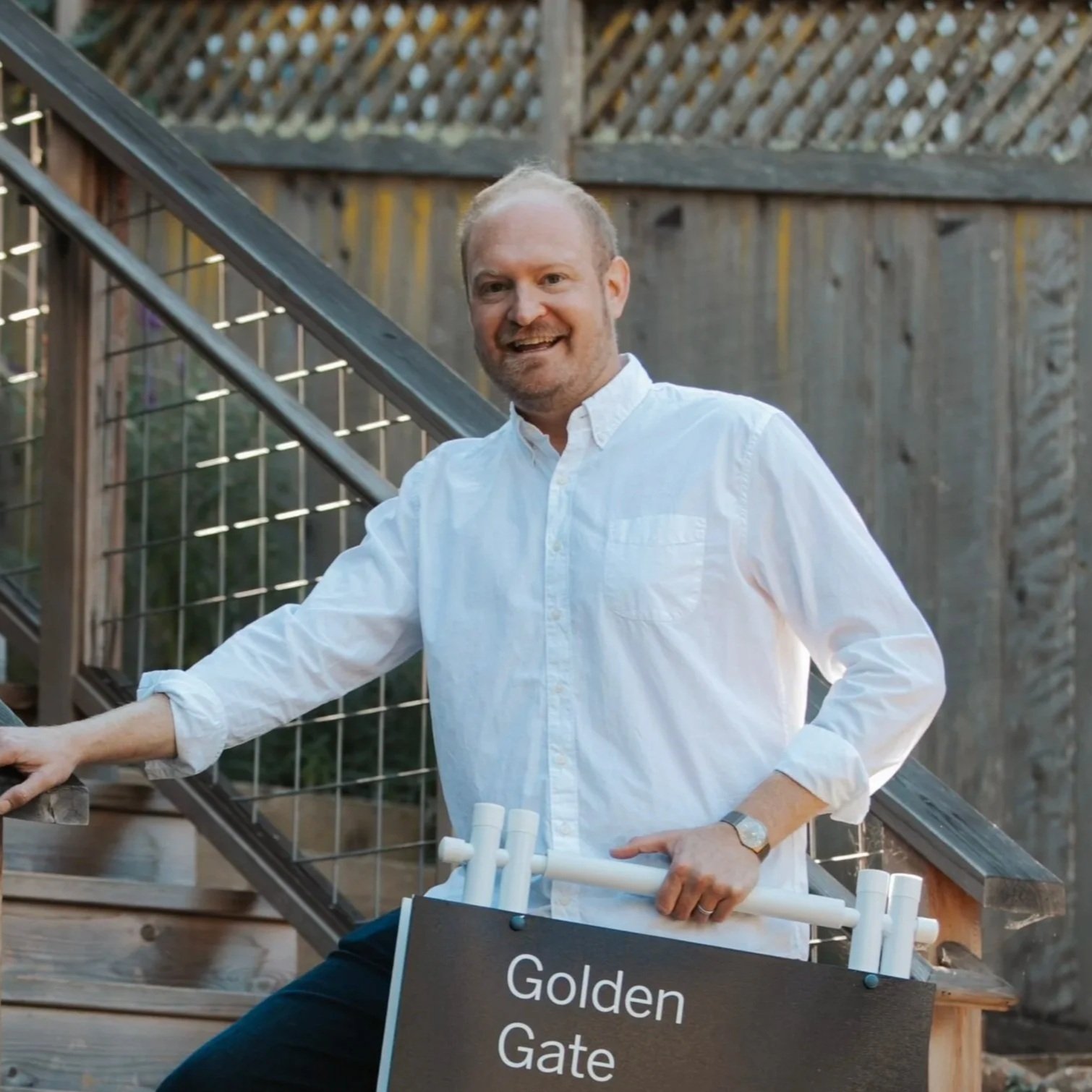 A man in a white button-up shirt with rolled sleeves, smiling, sits near a sign that says "Golden Gate." He is outdoors near a staircase with wooden railings and a wire fence, with a wooden fence in the background.