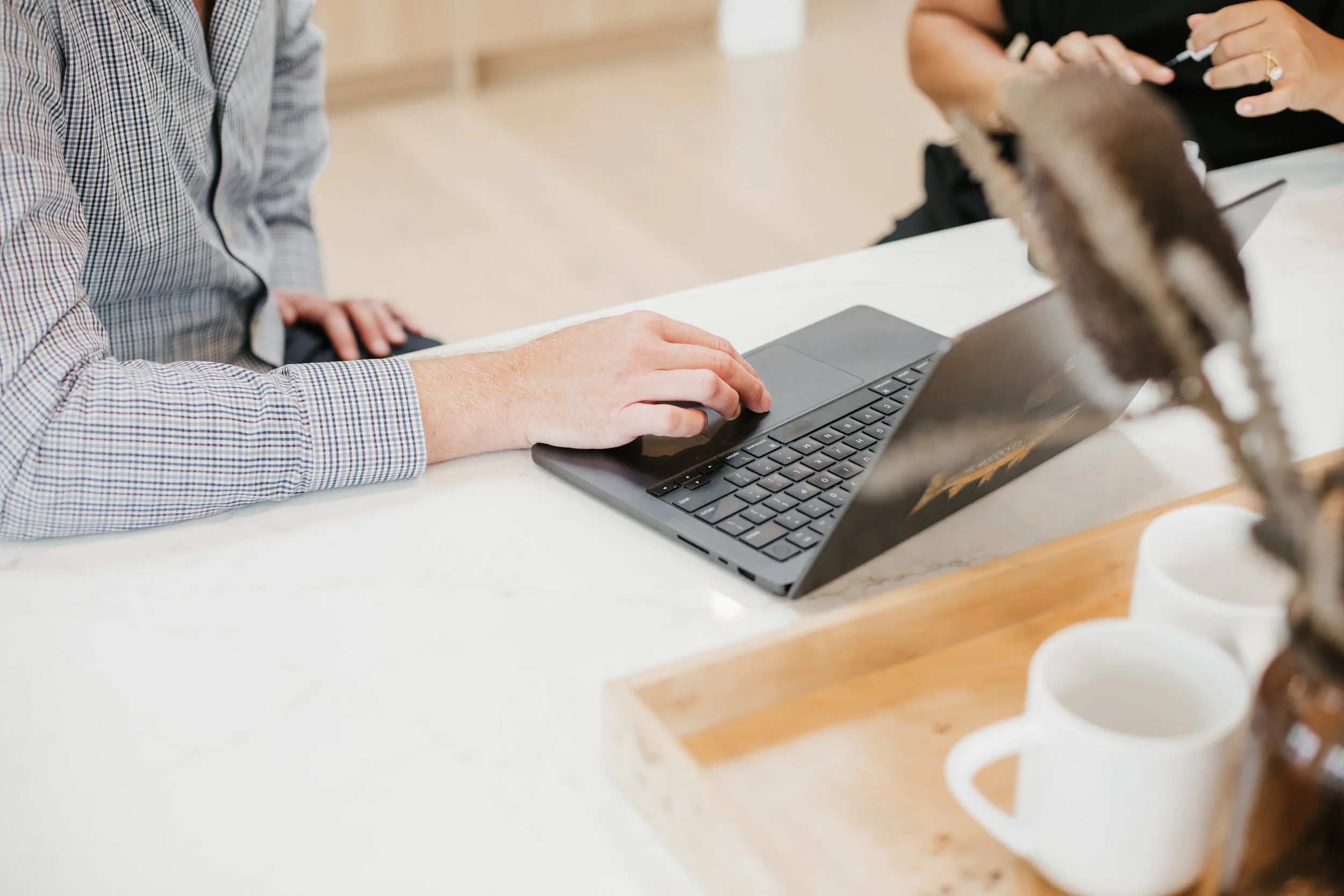 Two people sitting at a table working on a black laptop. One person wears a checkered shirt; the other has a ring on their finger. There are white mugs in a wooden tray in the foreground.