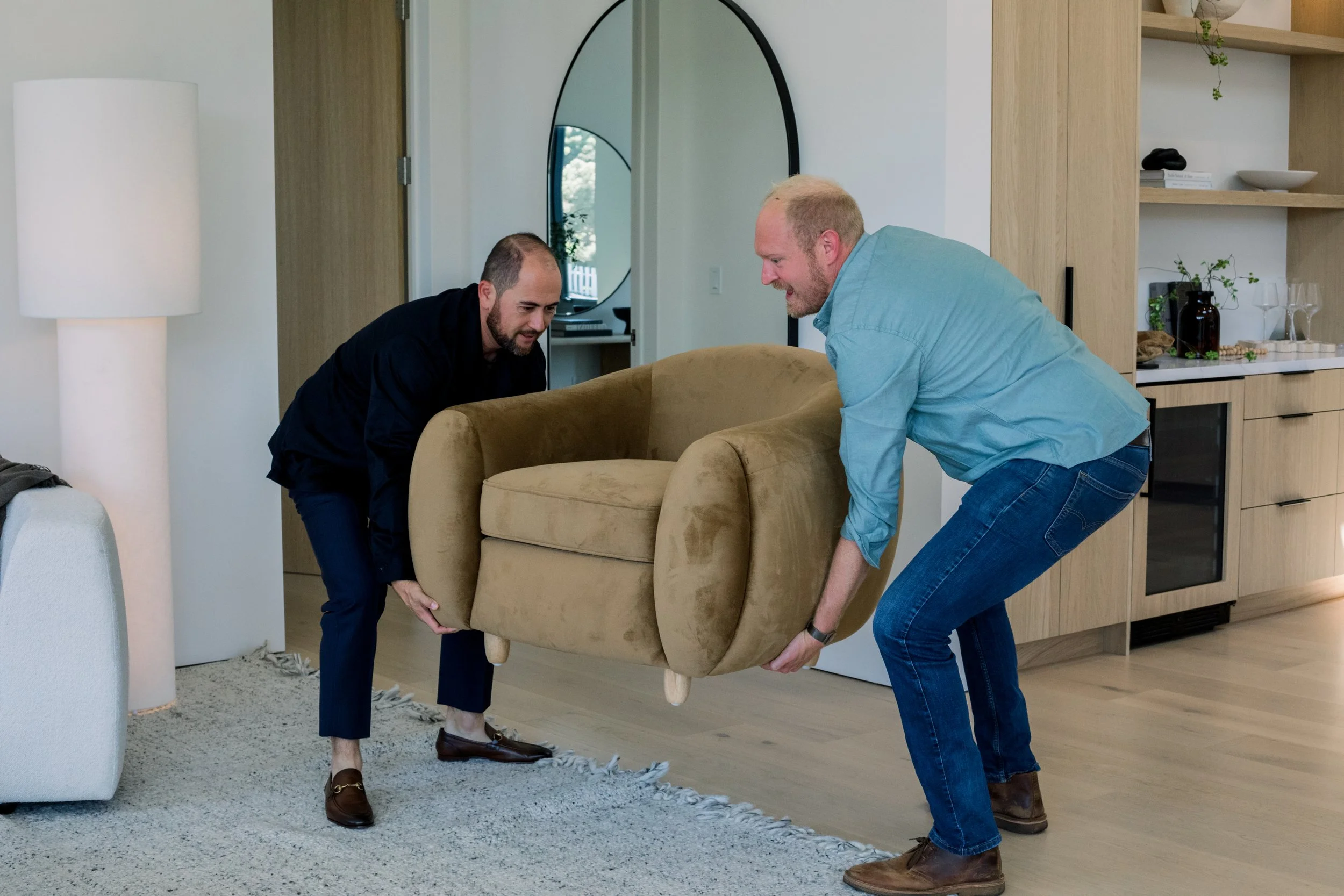 Two men are lifting a beige armchair together inside a modern living room. They are smiling and working as a team.