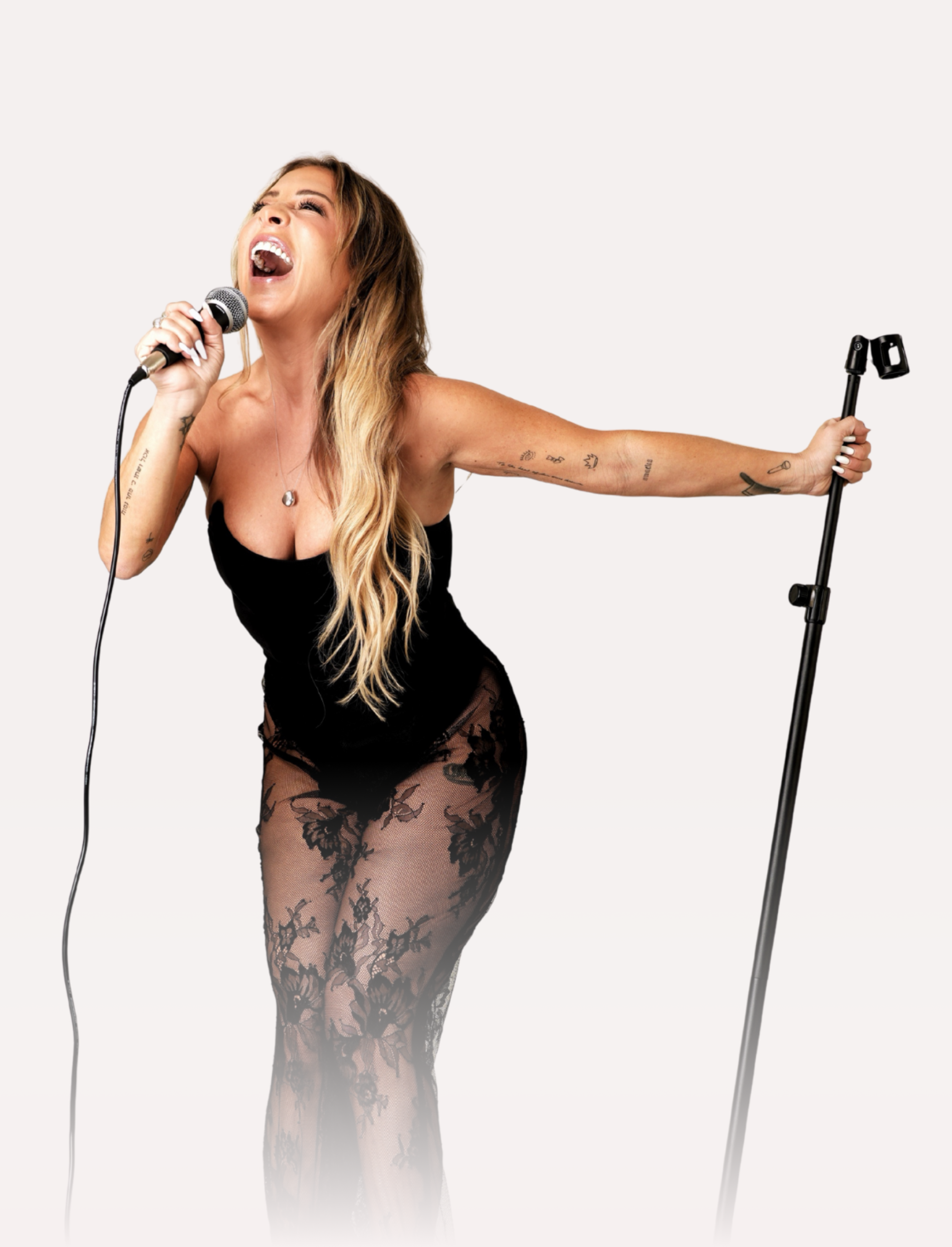 A woman speaking passionately into a microphone, wearing a black outfit with lace tights, holding a microphone stand, with tattoos on her arms, and mid-performance expression.