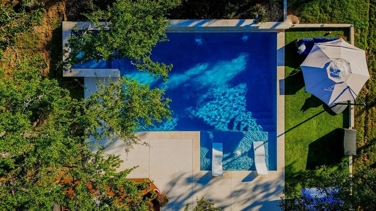 Aerial view of a swimming pool with chairs at the poolside, an umbrella, and surrounding greenery.