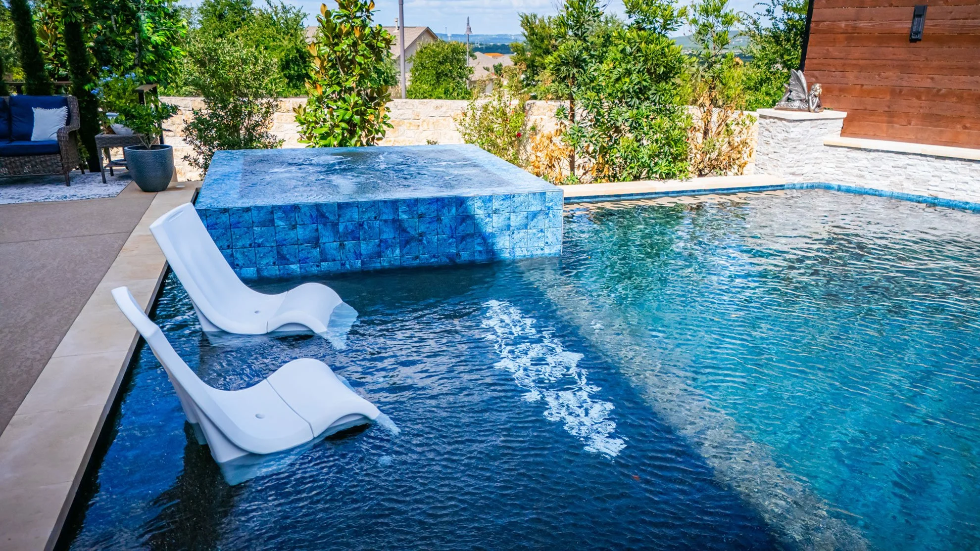 A backyard pool with two white lounge chairs partially submerged in water, a raised blue-tiled spa, lush green trees and bushes, an outdoor seating area with a wicker couch, and a wooden wall with a decorative sculpture in the background.