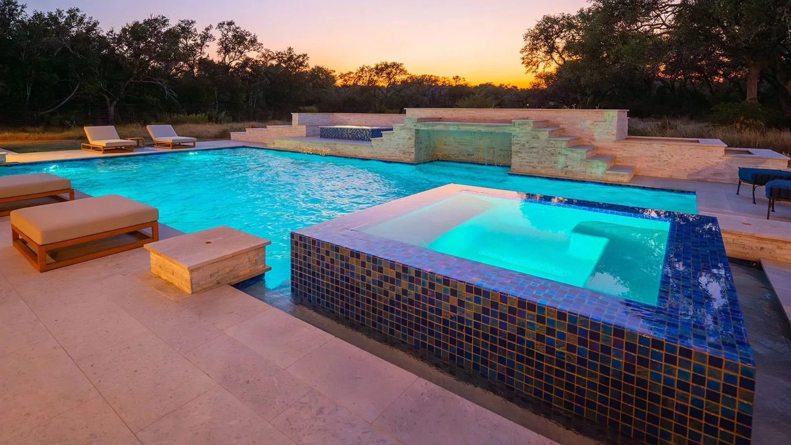 Luxury backyard pool with a hot tub, outdoor loungers, and a waterfall feature during sunset.