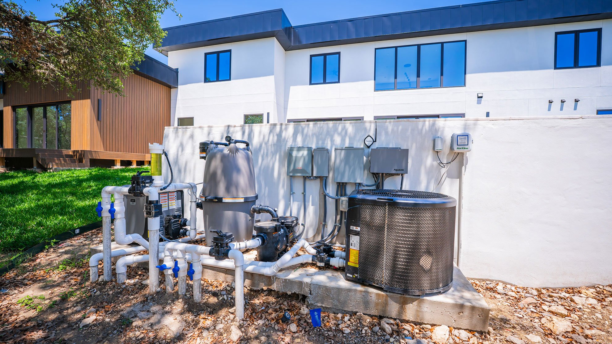 Outdoor view of a modern building behind a white wall with an installed pool equipment system, including filters, pumps, and control panels, on a concrete platform, surrounded by grass and trees.