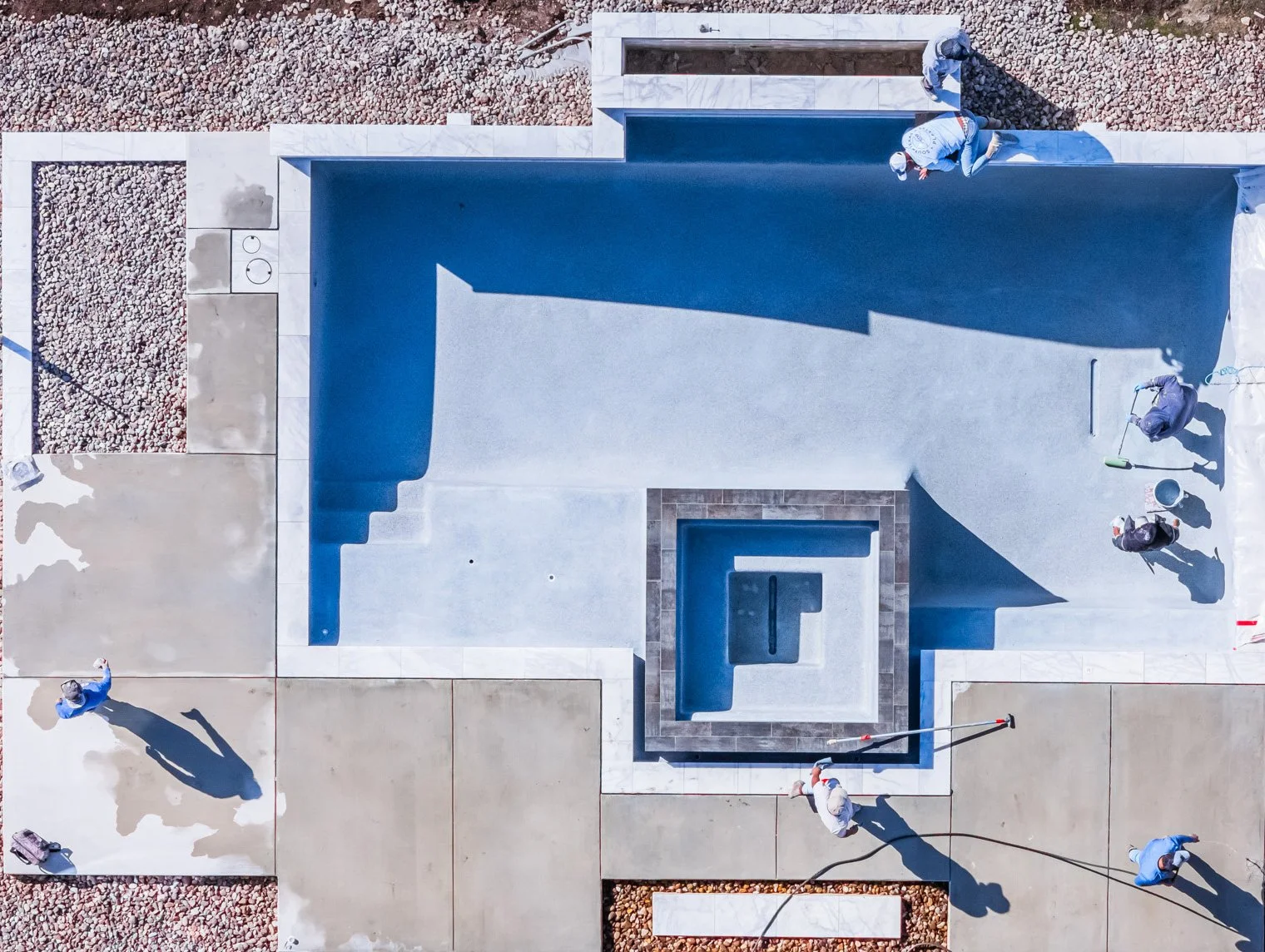 An aerial view of a construction site with workers installing a swimming pool. The pool is partially filled and surrounded by concrete and tile surfaces. Gravel is visible around the site, and workers are using tools such as rollers and hoses.