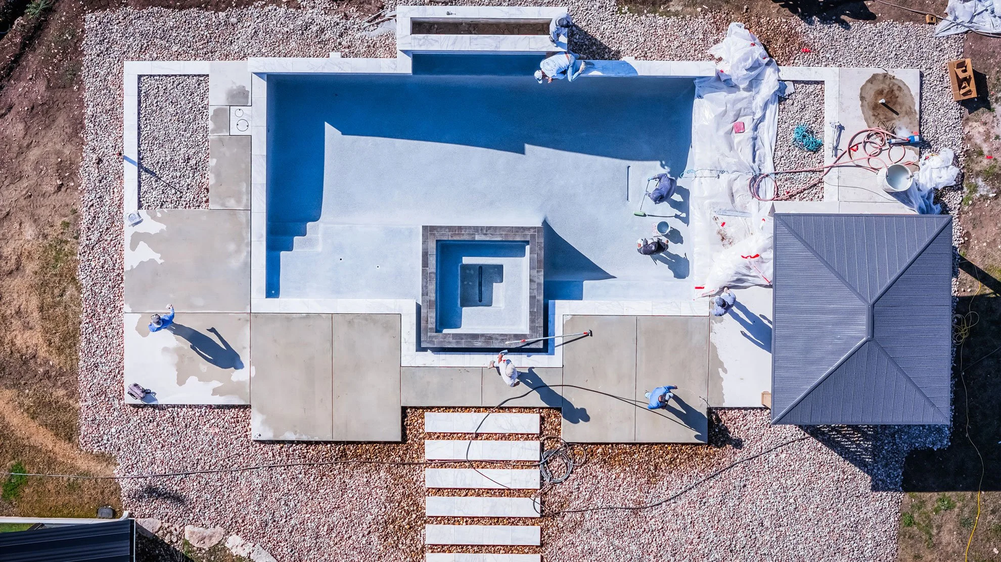 An aerial view of a swimming pool under construction, with workers and construction materials around the area. The pool is partially filled with water.