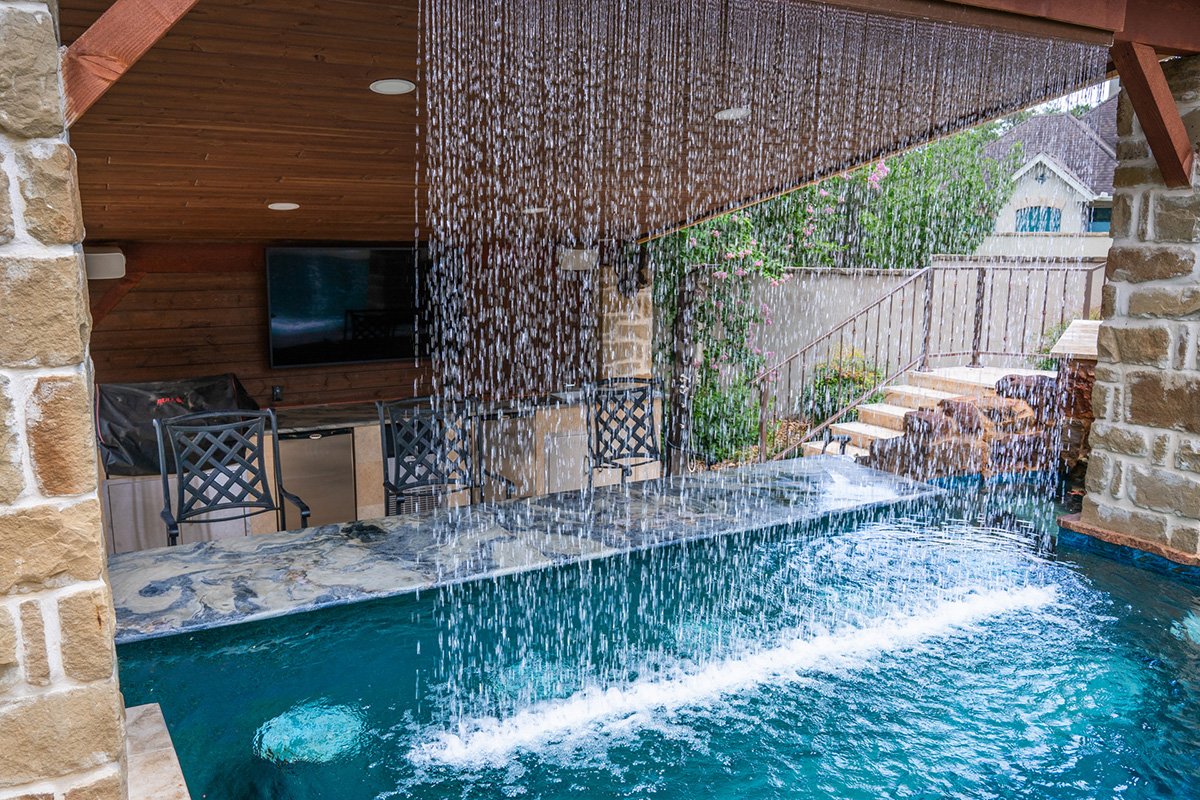 Indoor pool with a waterfall feature cascading into the water, surrounded by brick and wooden accents, with outdoor stairs, garden, and residential houses visible in the background.
