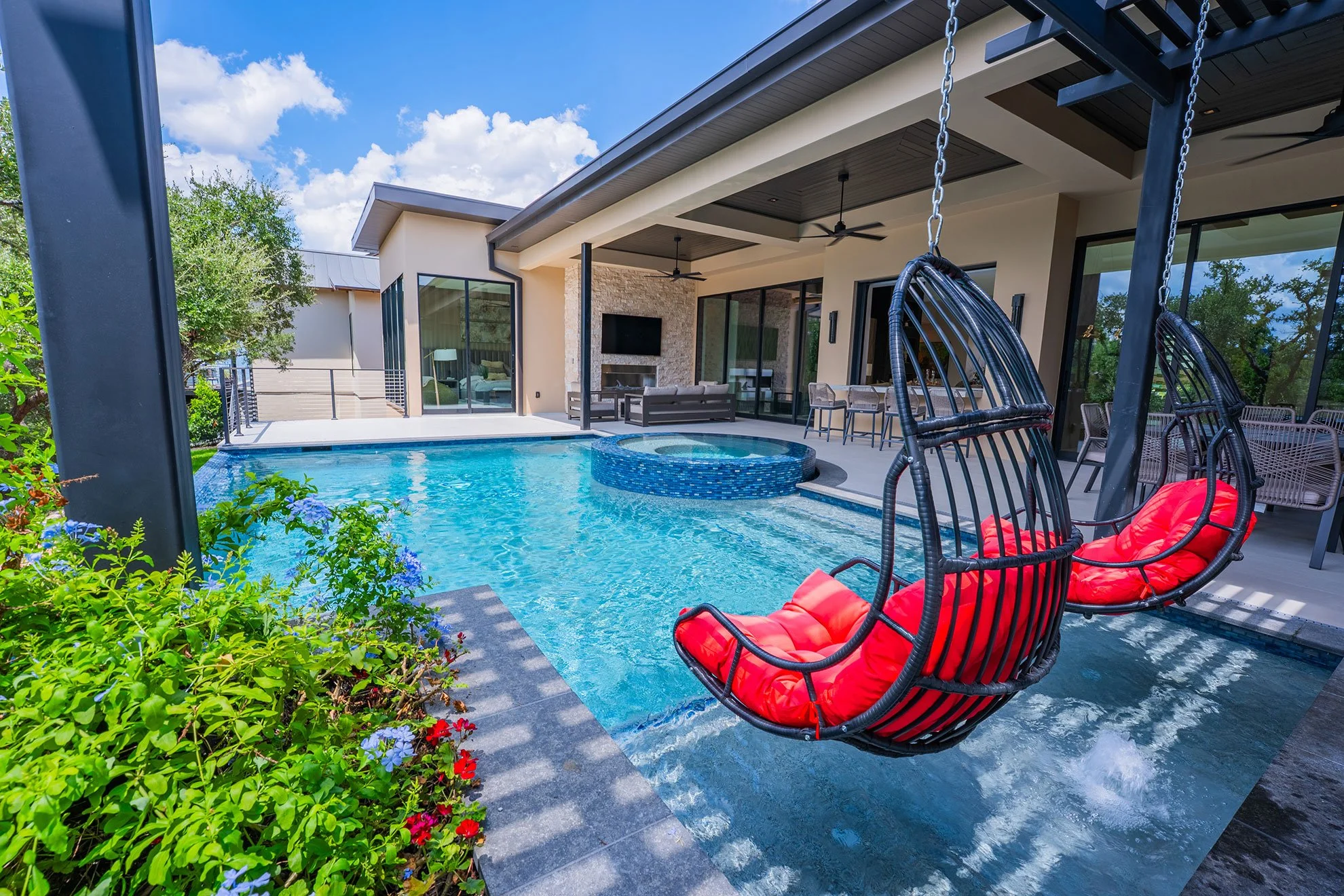 Modern backyard with a swimming pool, two hanging chairs with red cushions, a covered patio with outdoor furniture, and a house with large glass sliding doors under a blue sky with clouds.
