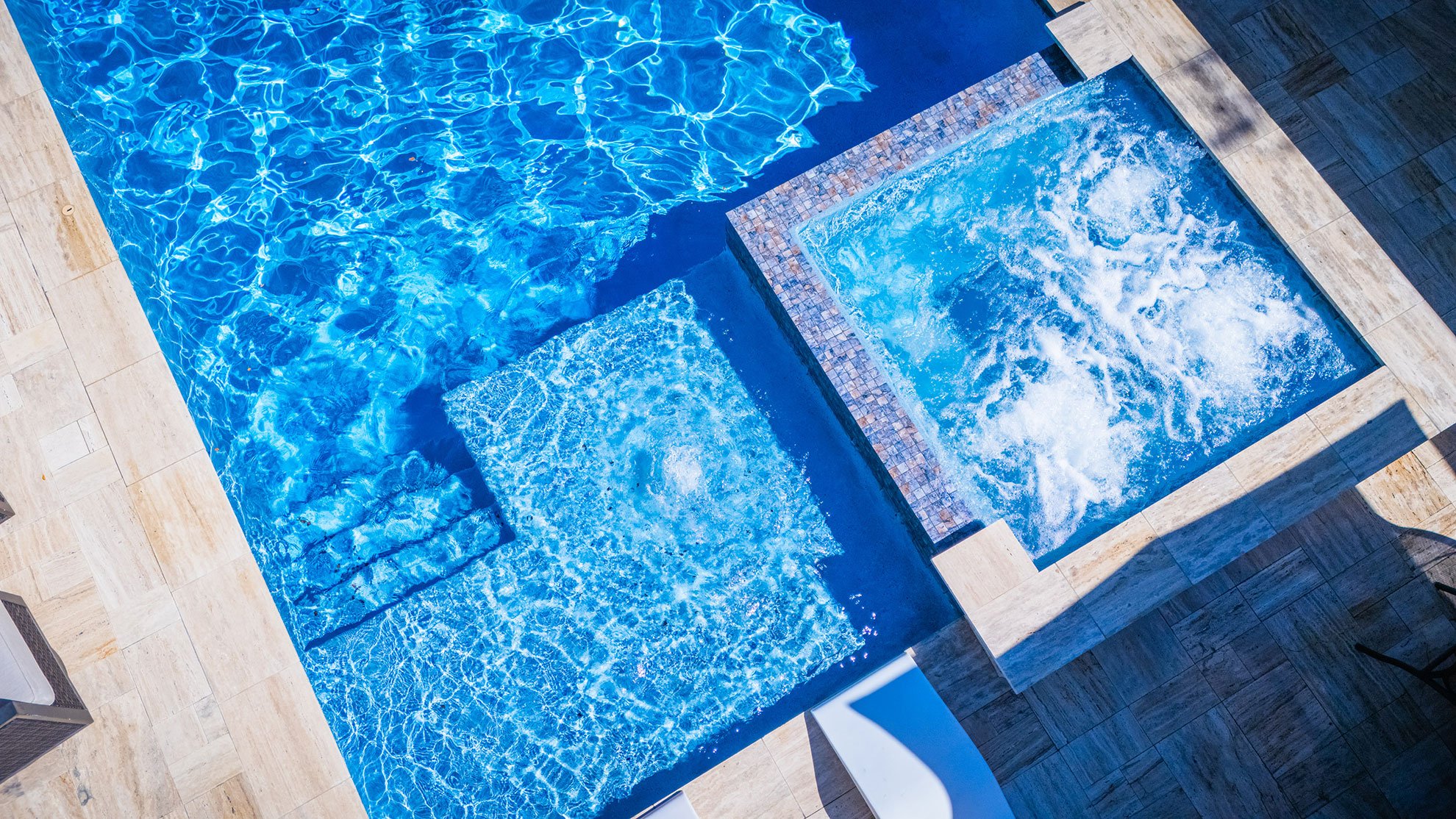 Overhead view of a swimming pool with two hot tubs, one bubbling and the other calm, surrounded by tiled flooring and furniture.