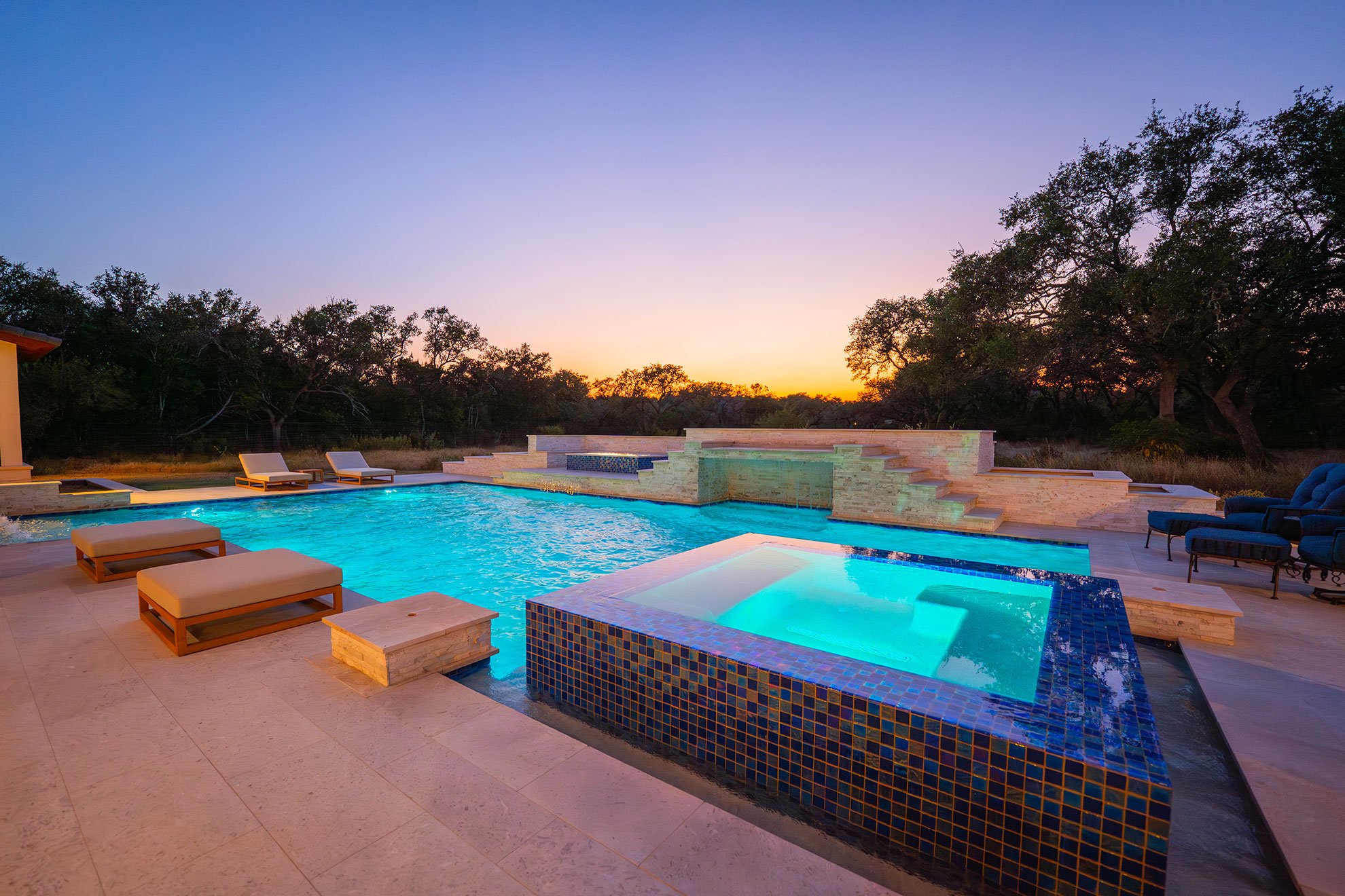 Luxury swimming pool with lounge chairs, a hot tub, and a sunset view behind trees.