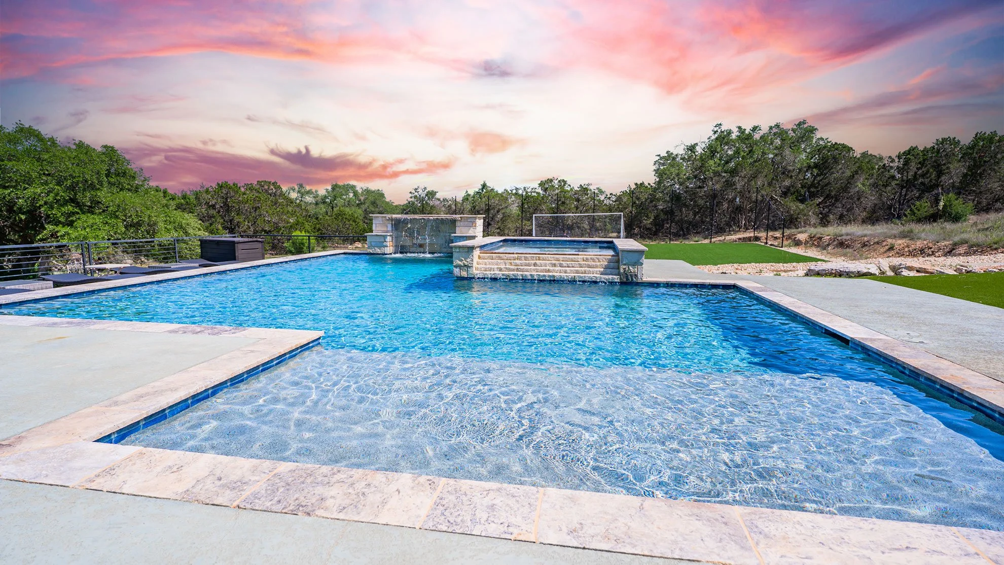 An outdoor swimming pool with a cascading waterfall feature, surrounded by a concrete deck and greenery, under a pink and purple sunset sky.