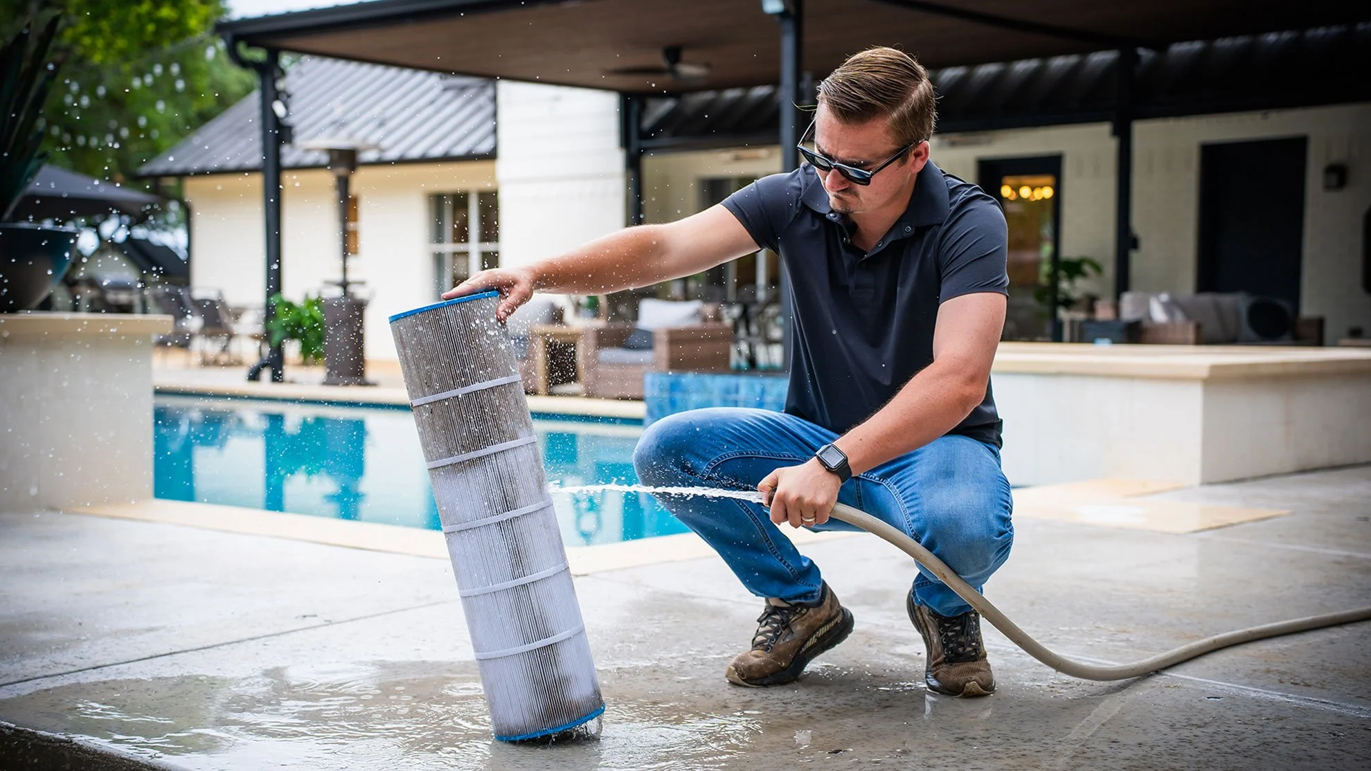 A man wearing sunglasses, a dark polo shirt, and jeans crouches while cleaning a pool filter with a garden hose near a backyard pool.