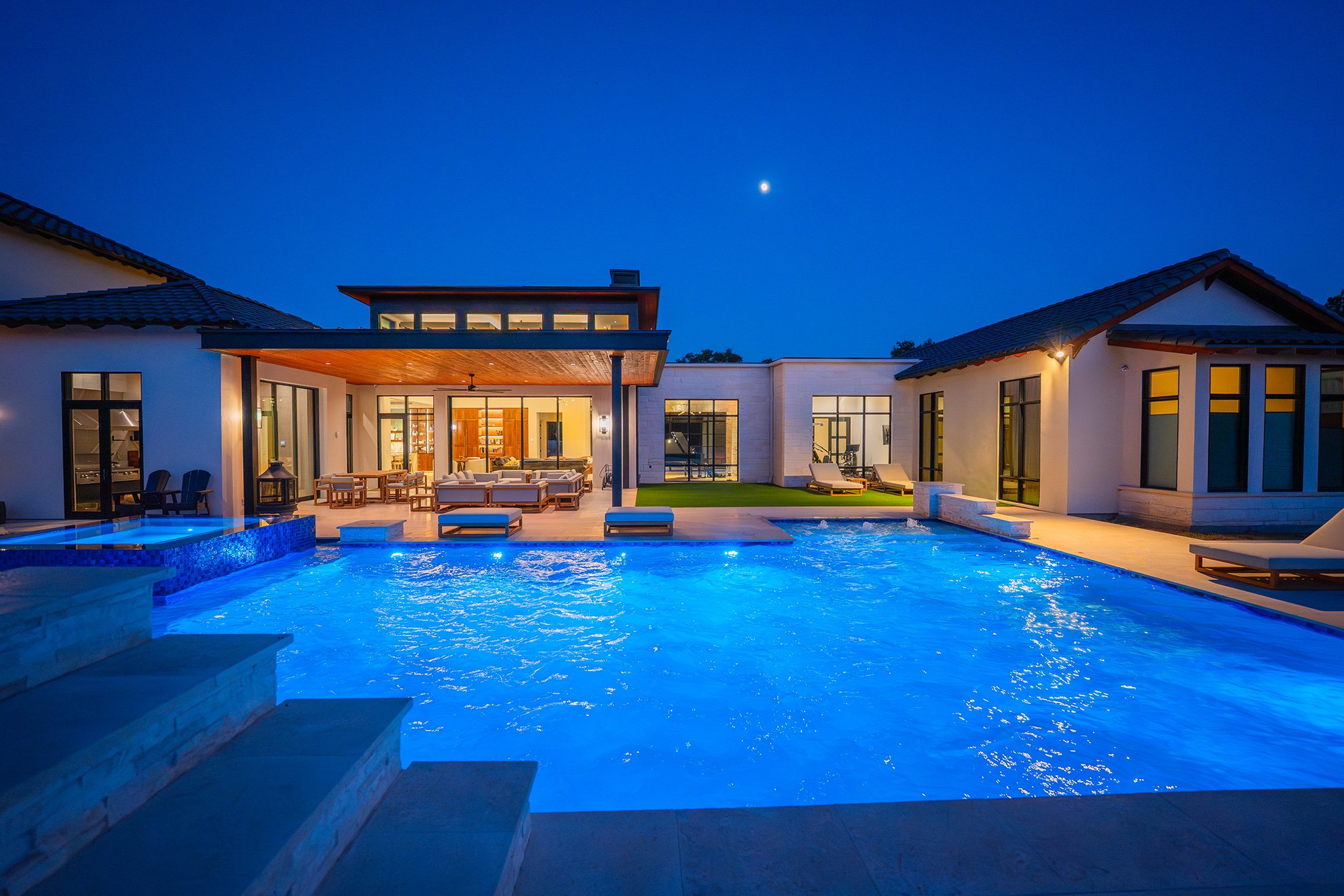 A modern house with a swimming pool in the backyard during evening, with the moon visible in the sky and warm interior lights.