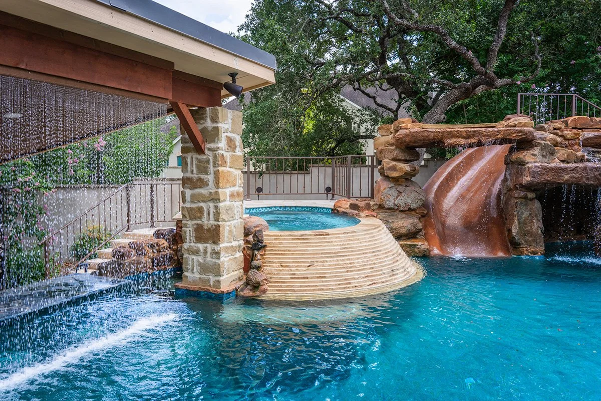A swimming pool with a built-in hot tub, surrounded by rocks and a waterfall feature, with trees in the background and a fence around the area.