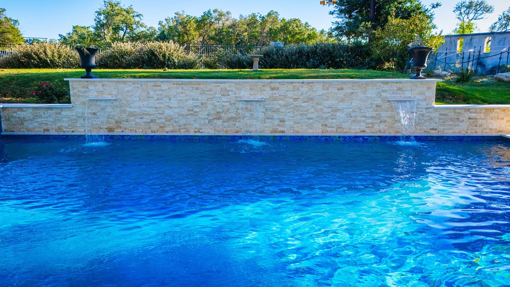 A rectangular blue swimming pool with a stone wall fountain feature that has three streams of water flowing from it. The background shows a grassy area with trees, shrubs, and decorative urns on top of the stone wall.