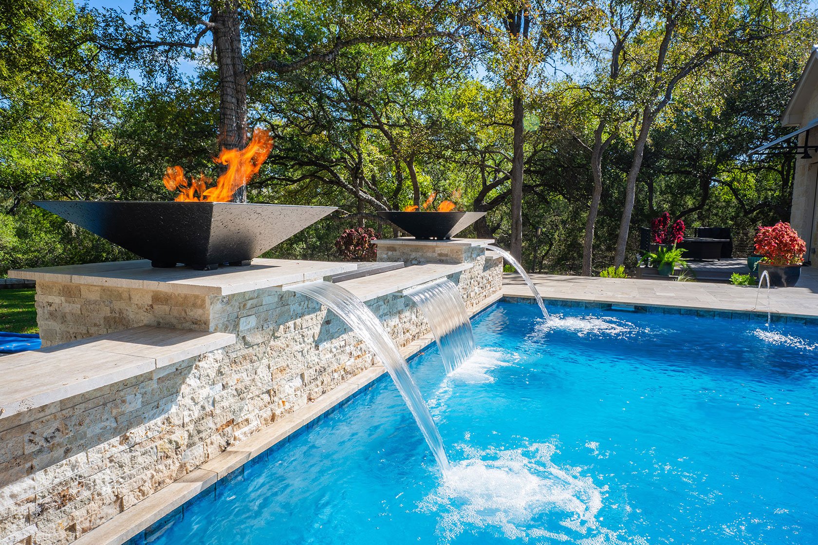 A backyard swimming pool with a stone edge, featuring three water spouts and two fire bowls with flames, surrounded by trees and potted plants.