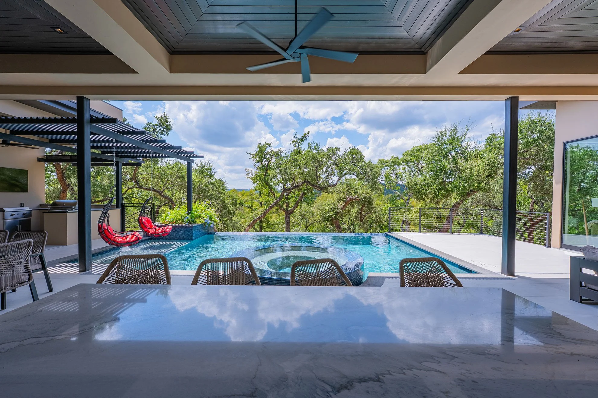 Modern outdoor living area with a pool, slide, and surrounded by green trees under a partly cloudy sky.