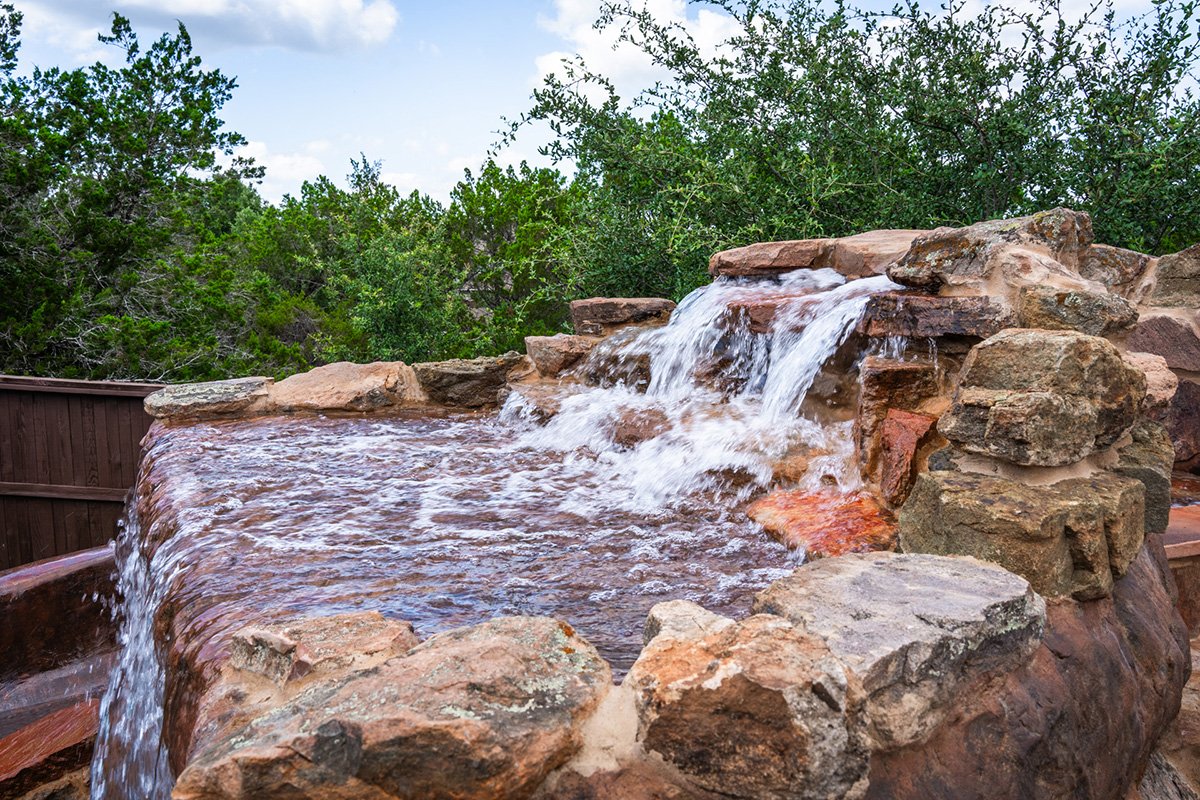Water feature with a small waterfall flowing over rocks, surrounded by green trees under a partly cloudy sky.