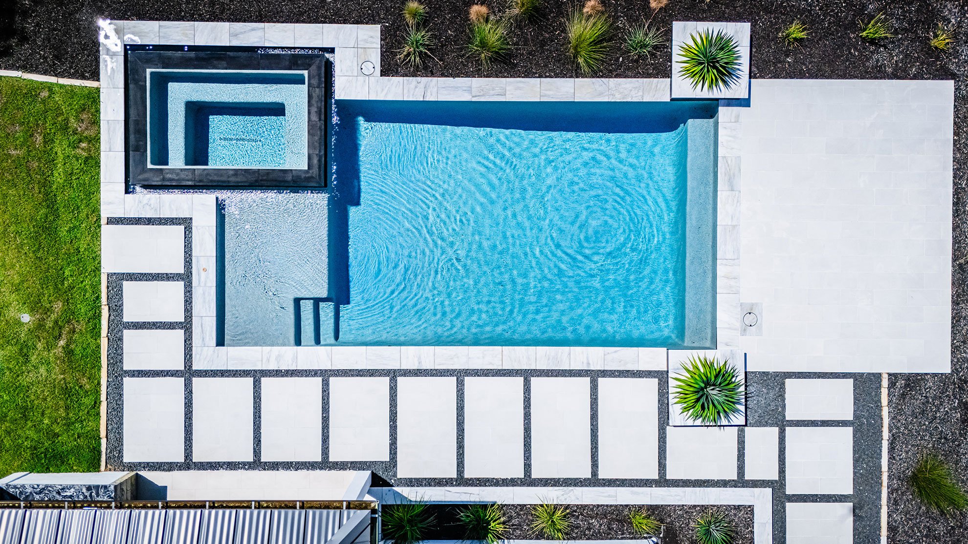 Aerial view of a modern swimming pool with a small raised hot tub or spa at one end, surrounded by white concrete tiles, green plants, and landscaped areas.