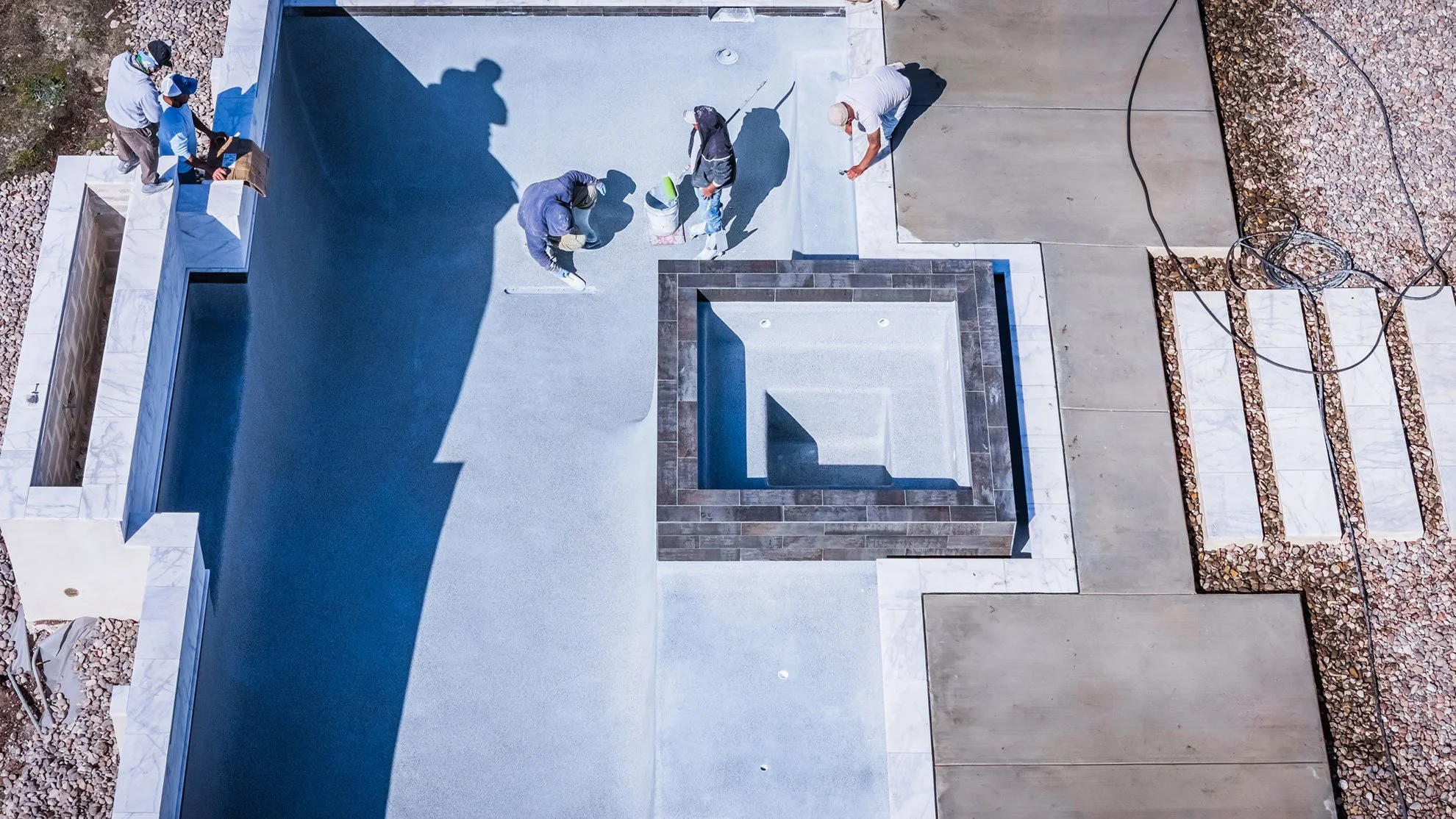 Construction workers installing a flat roof with white coating, an empty square pool with brown tiles, and surrounding concrete and stone areas.