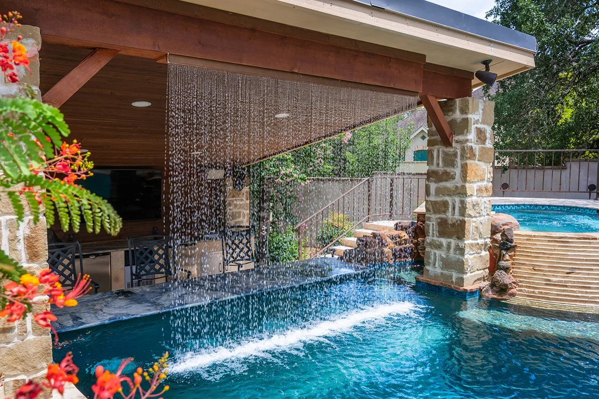 A backyard swimming pool with a waterfall feature and a shaded patio area, surrounded by greenery and fencing.