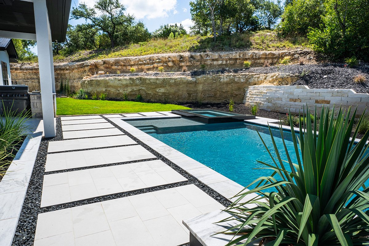 Backyard with a modern swimming pool, white stone pathway, green grass, and a rocky hillside in the background.