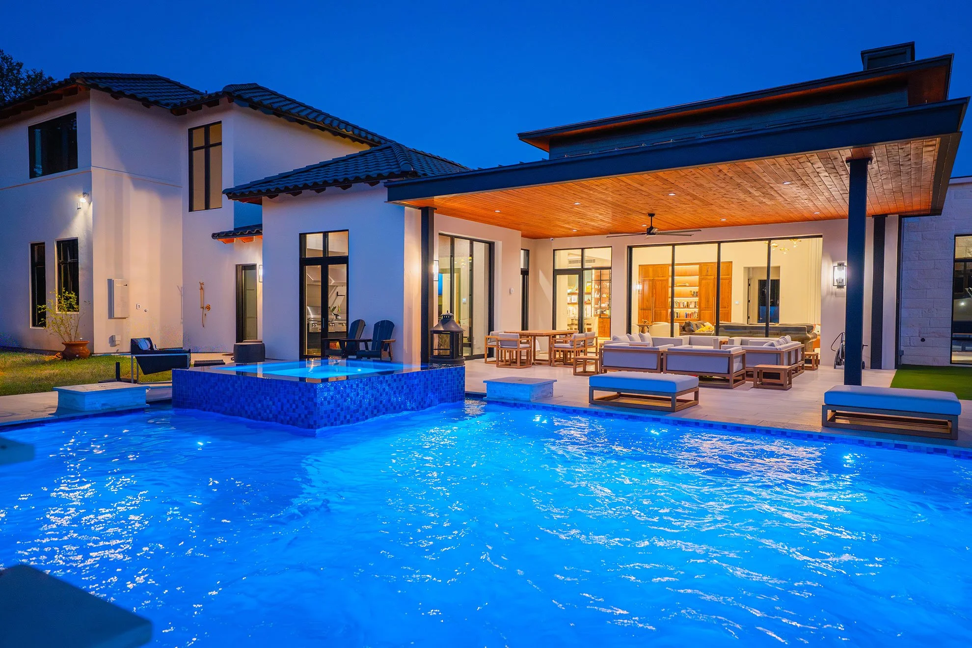 Luxury house with a lit backyard pool, outdoor lounge area, and open indoor living space visible through large glass doors at night.