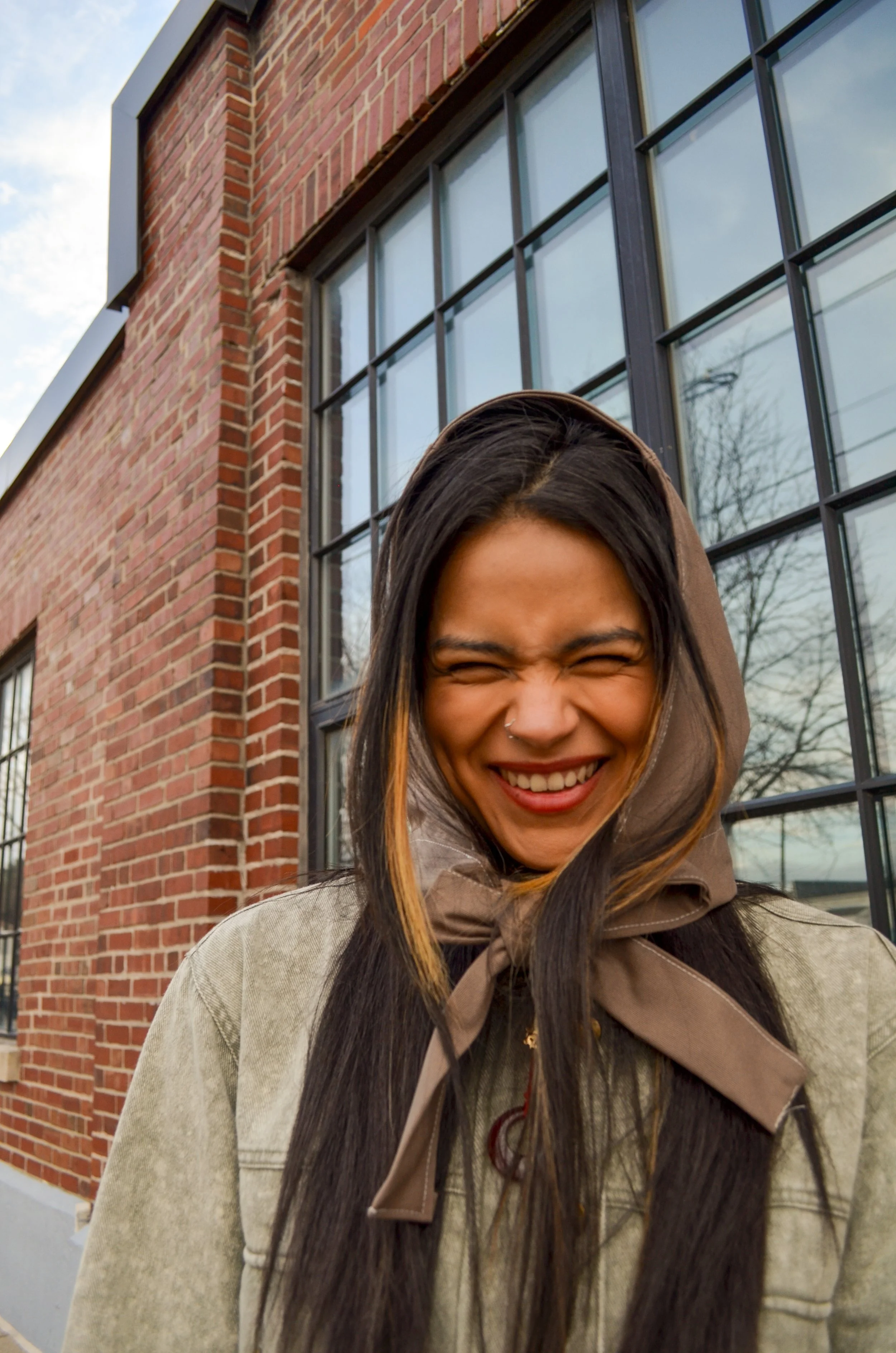 A young woman smiling with eyes closed outside a brick building with large windows, wearing a beige jacket and a brown detatchable hooded scarf.