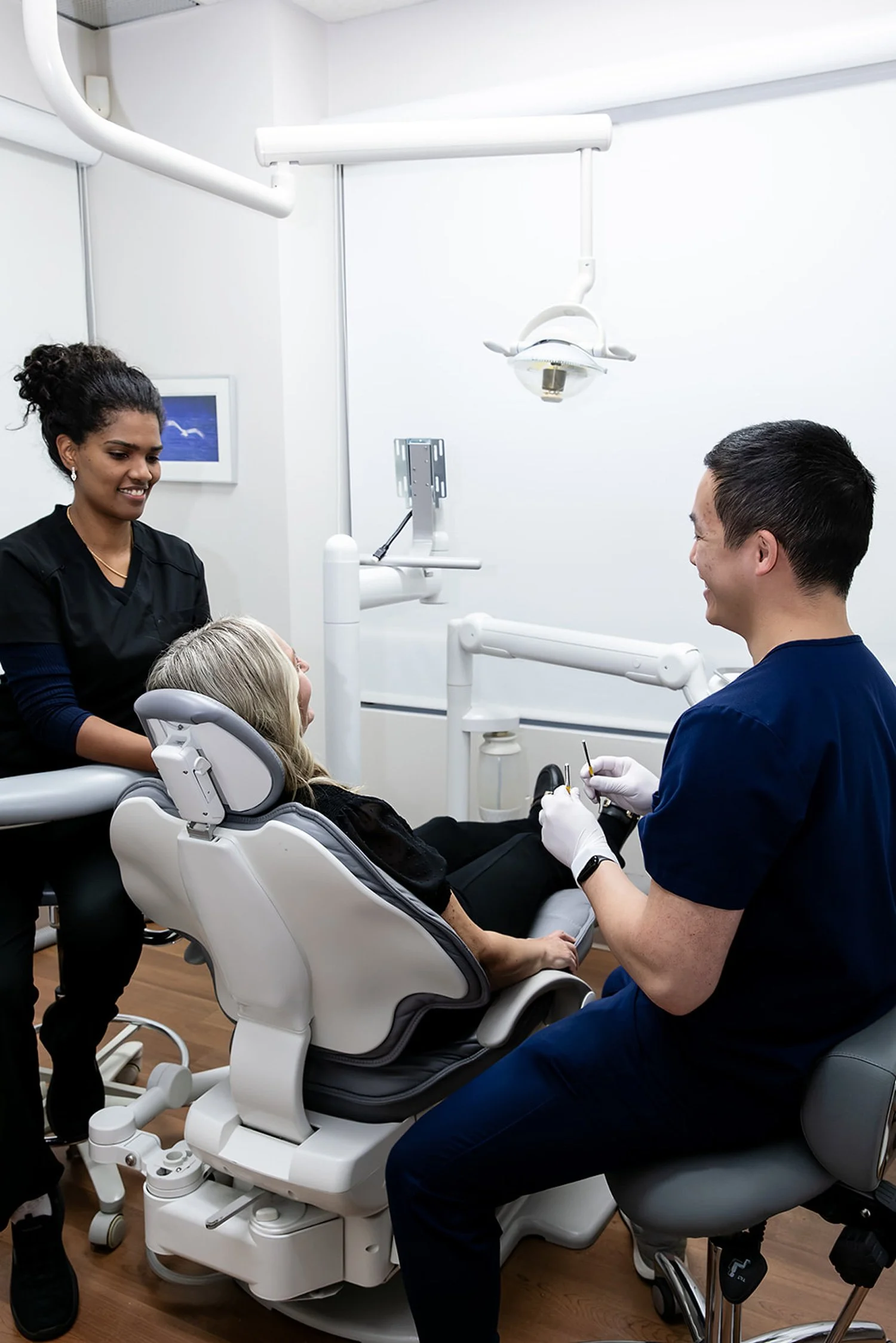 A woman is lying in a dental chair with two dental professionals attending her in a clean, modern dental office.
