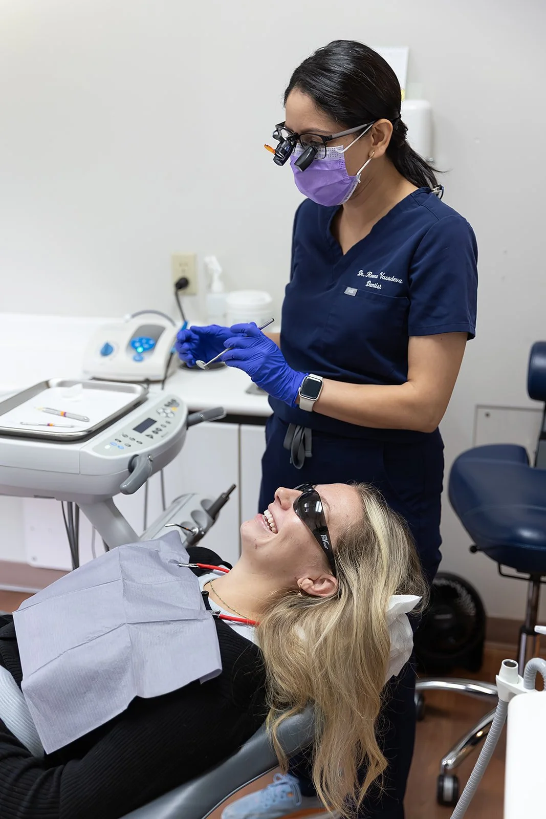 A woman lying in a dental chair smiling while wearing protective glasses, as a female dentist wearing gloves and magnifying glasses prepares dental tools.