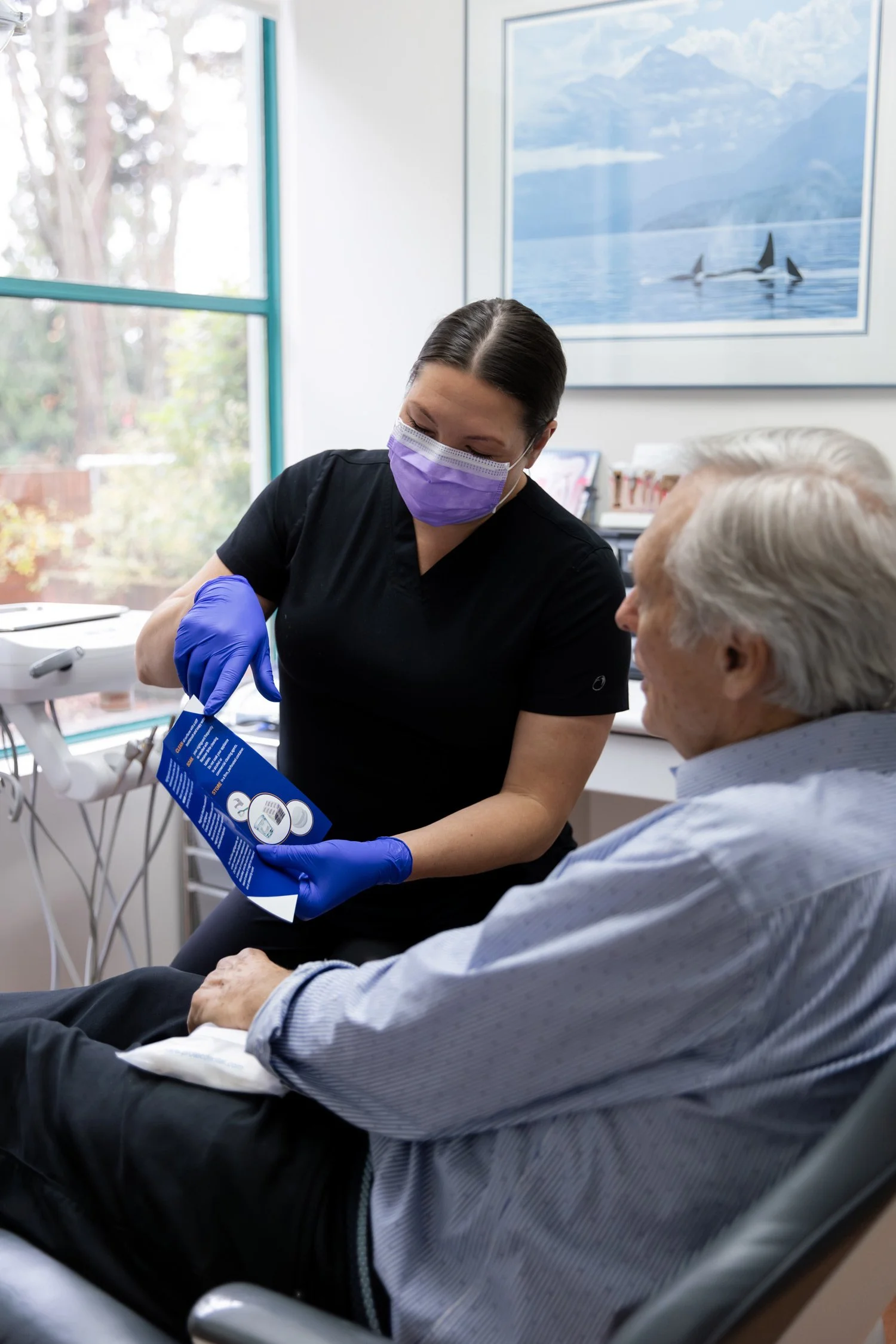 A healthcare worker wearing a purple mask and blue gloves showing a medical device package to an elderly man in a doctor's office.
