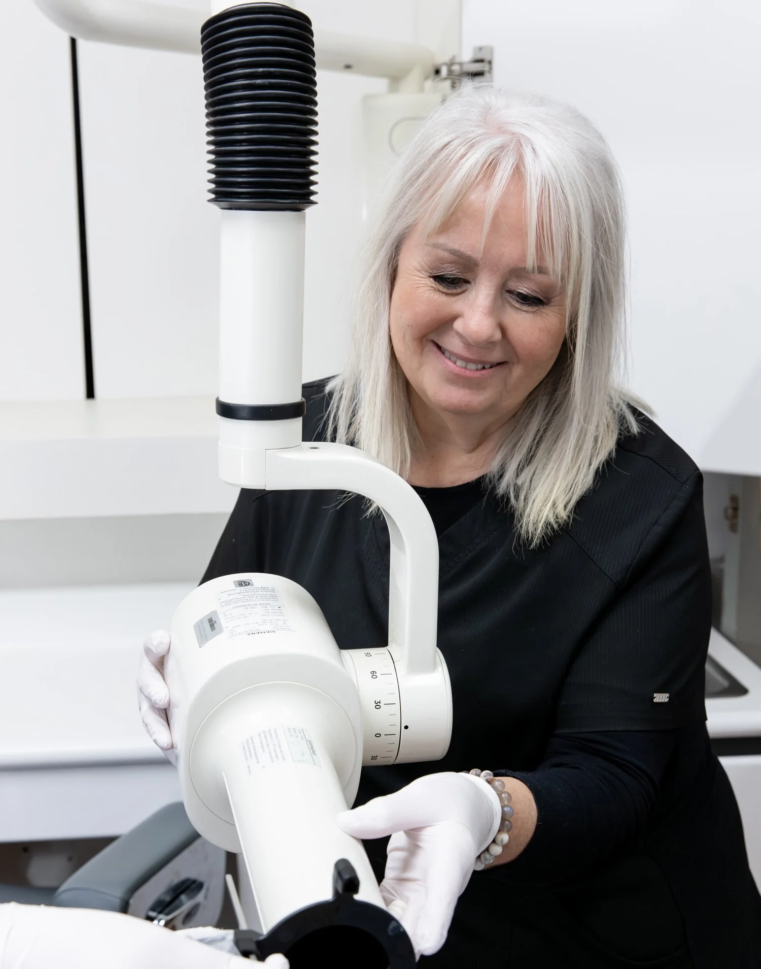 A woman with gray hair and wearing black scrubs and white gloves is looking down and smiling while using a medical or laboratory microscope in a white clinical or laboratory setting.