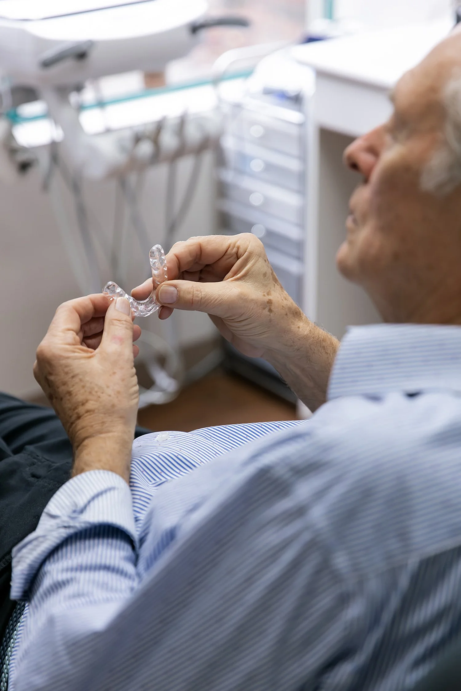 Senior man sitting in a dental chair holding a clear dental retainer, with dental tools and equipment in the background.