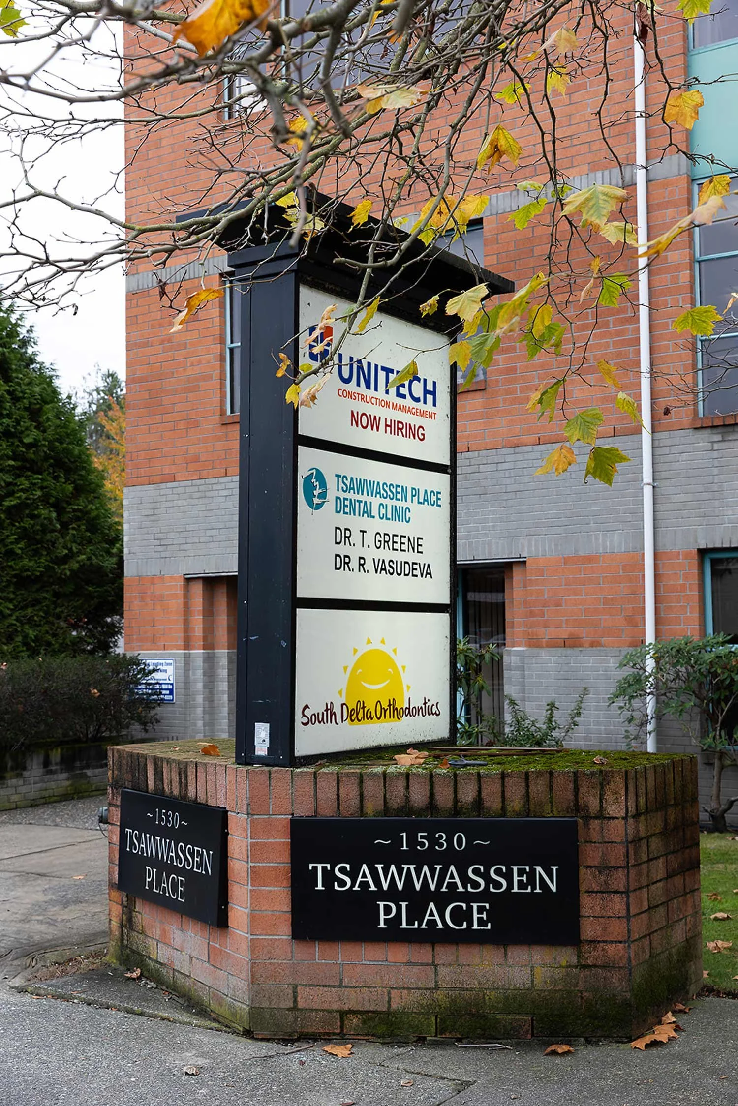 Sign on a brick and concrete base building listing tenants and businesses, including Unitech Construction Management, Tsawwassen Place Dental Clinic with Dr. T. Greene and Dr. R. Vasudeva, and South Delta Orthodontics, with signs on the building and a brick wall at the corner of Tsawwassen Place.