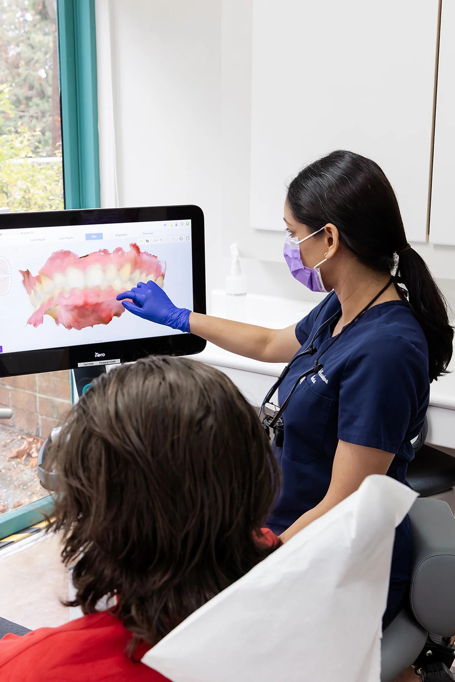 Dental professional wearing a purple mask and gloves showing a 3D digital image of teeth and gums on a computer screen to a patient in a dental office.